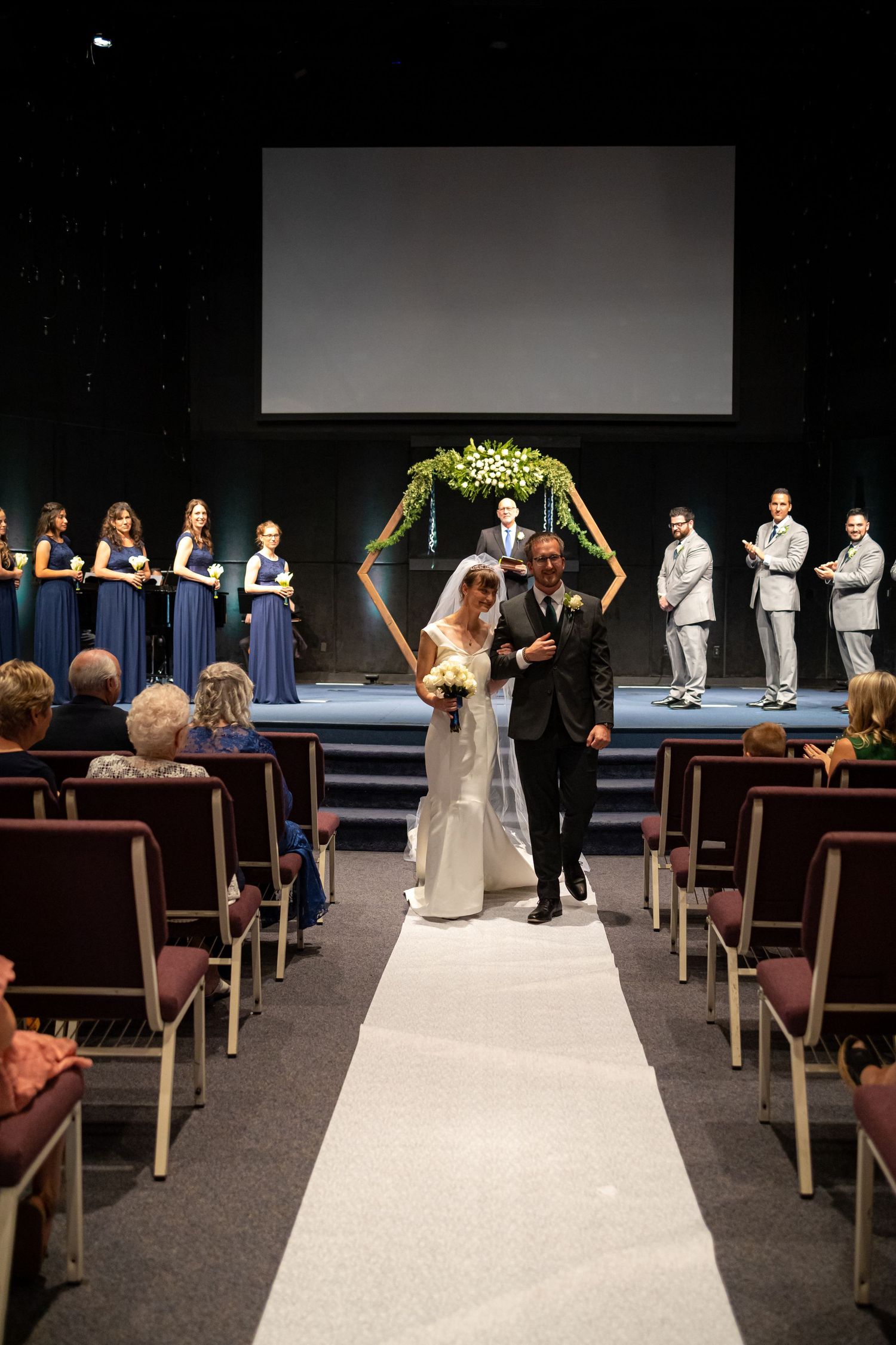 Couple walking down white aisle runner after ceremony with wedding party and guests looking on.