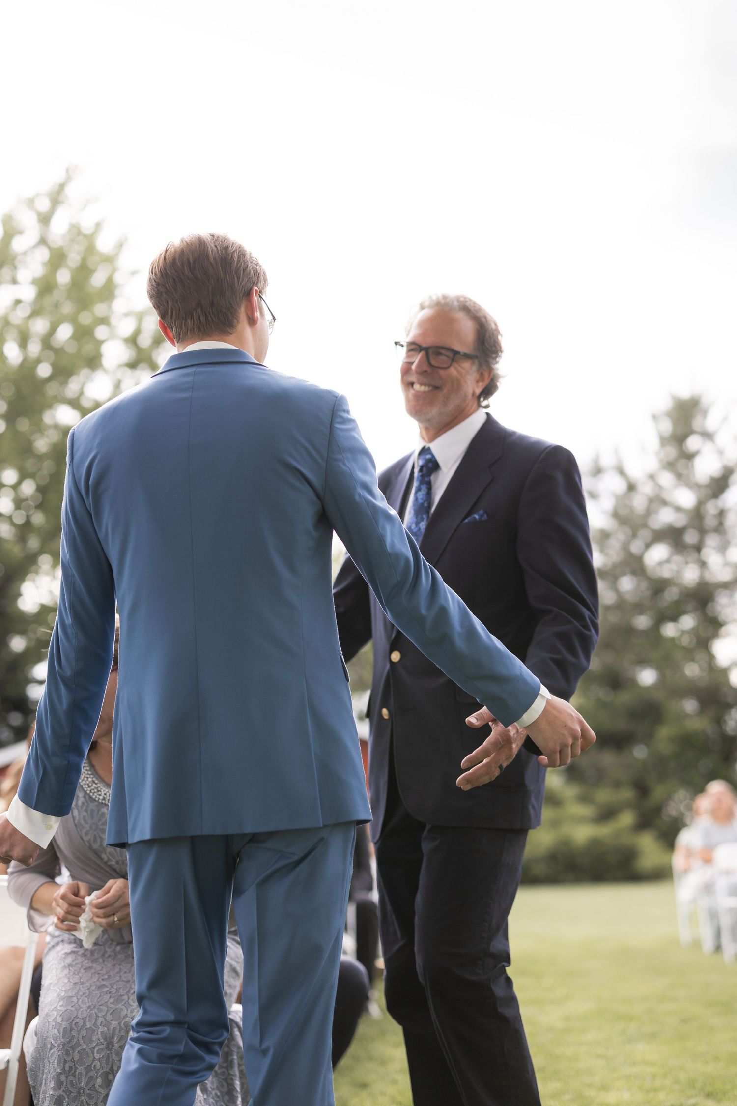 Two people in formal attire greeting each other at an outdoor wedding ceremony.