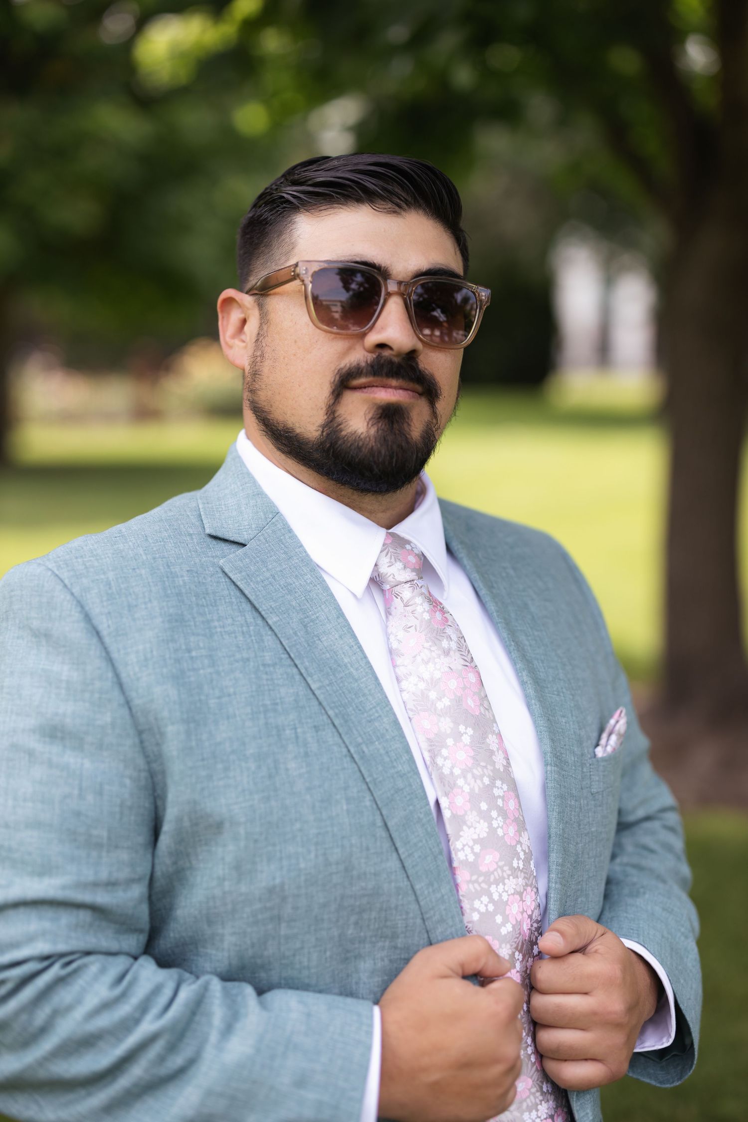 Portrait of a gentleman in light blue suit with floral tie and sunglasses outdoors.