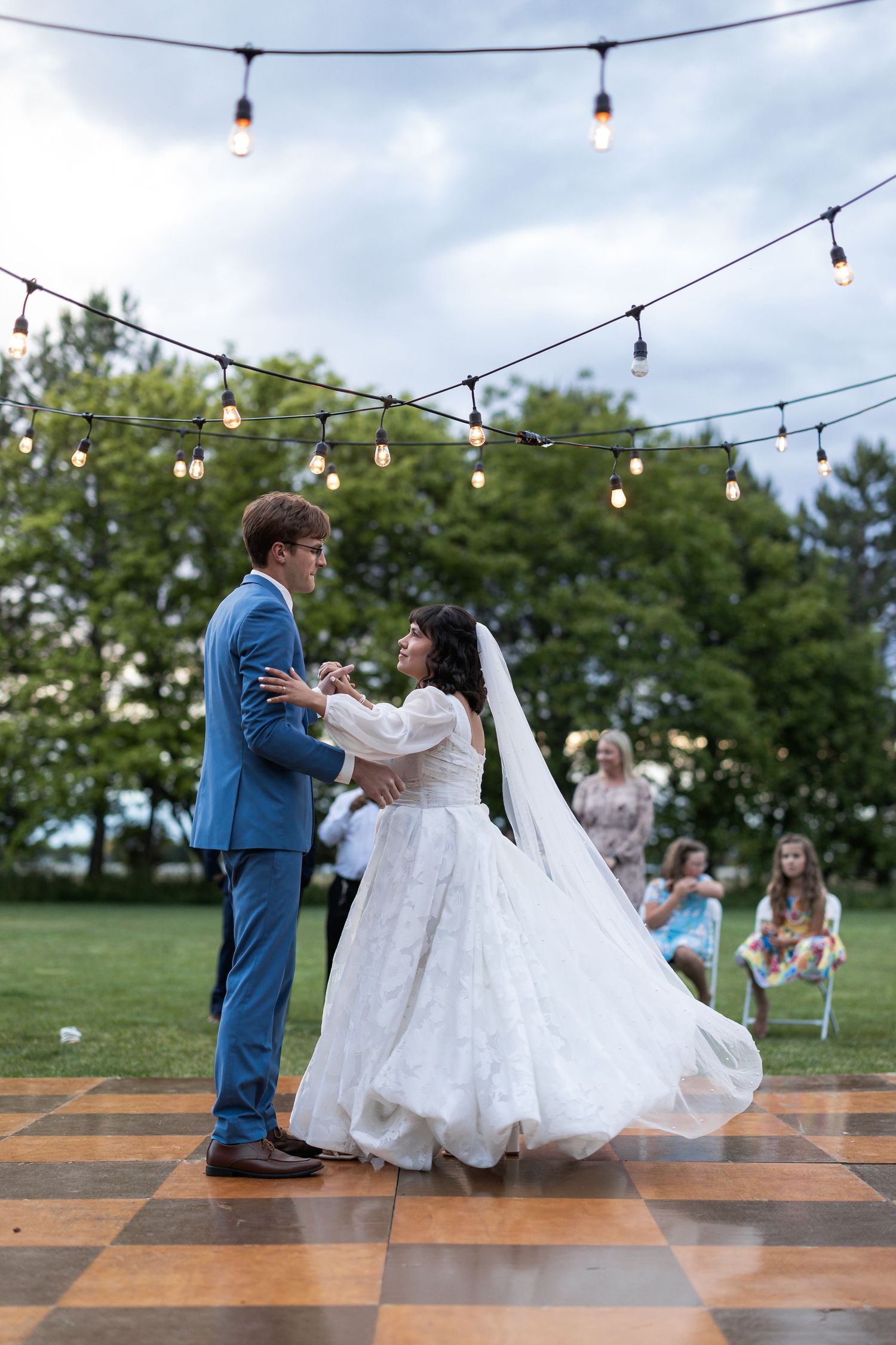 Newlyweds share first dance on outdoor checkered dance floor beneath string lights.