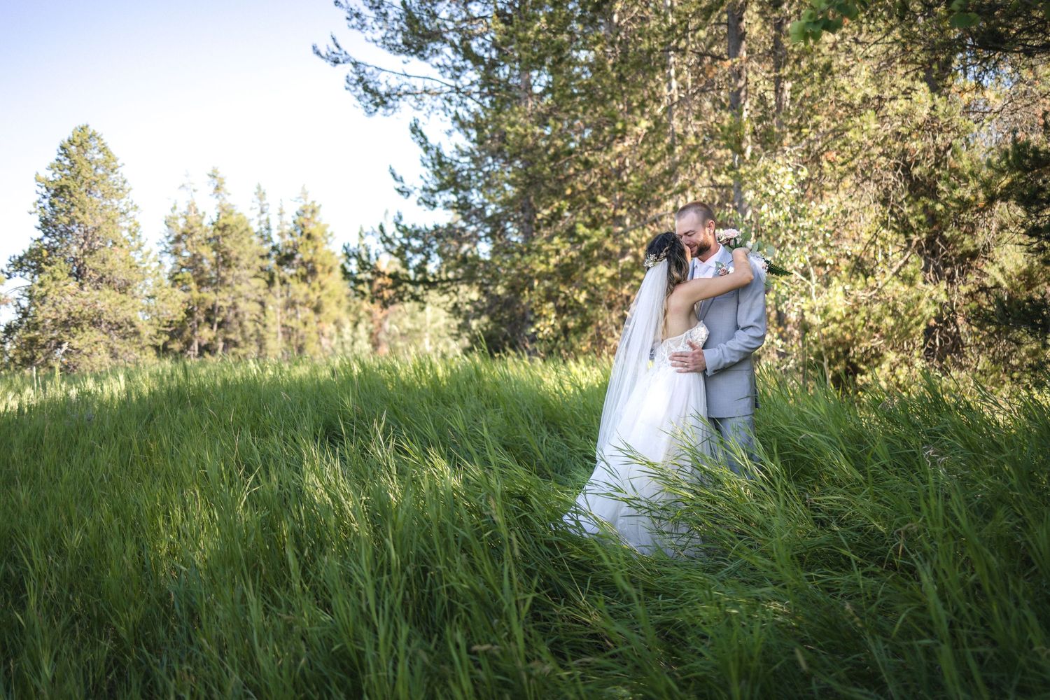 A wedding couple embraces in a sun-drenched field with evergreen trees in soft focus.