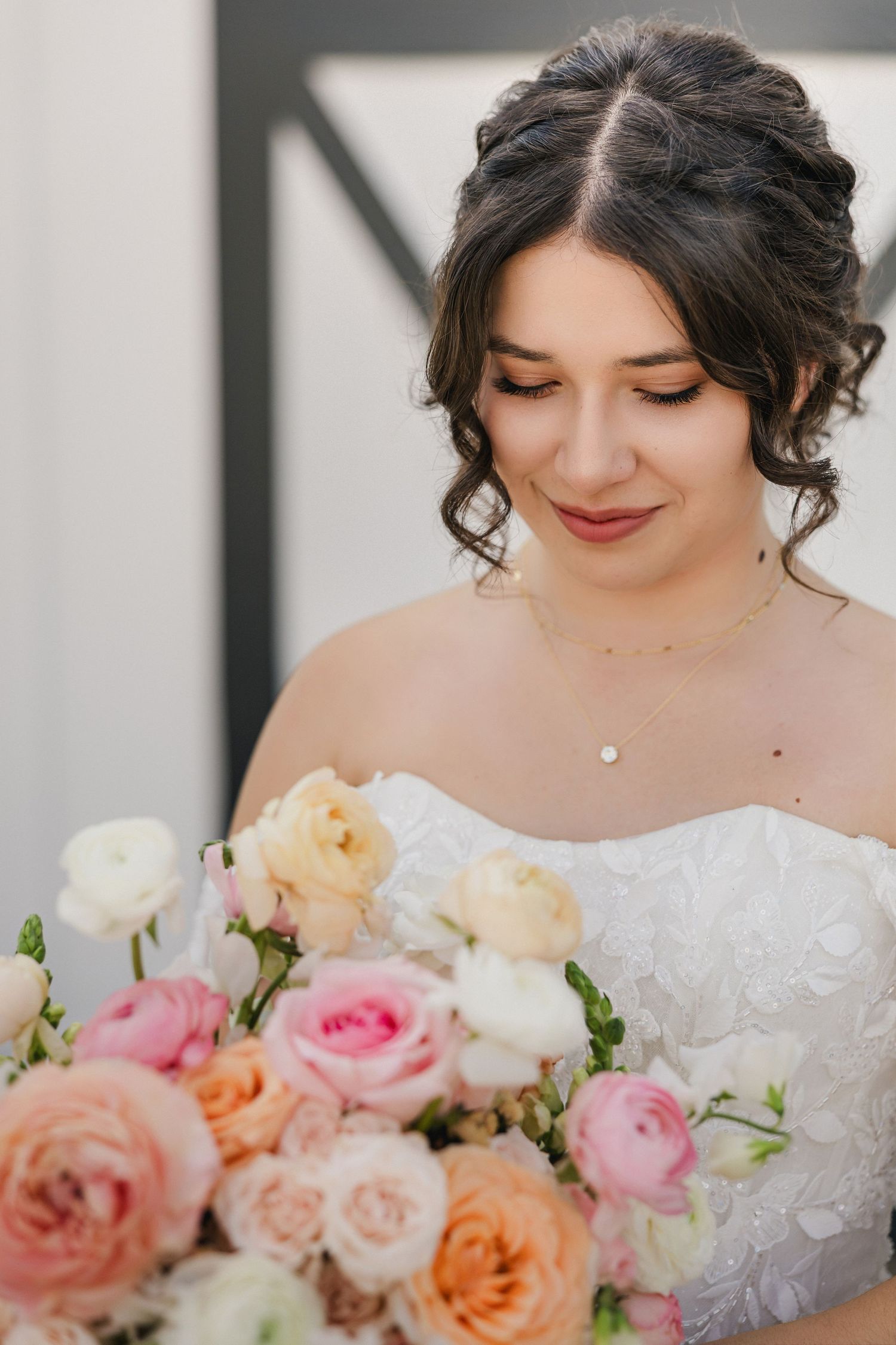 Bride in white strapless gown holds colorful bouquet of pink, peach, and white roses, smiling softly downward.