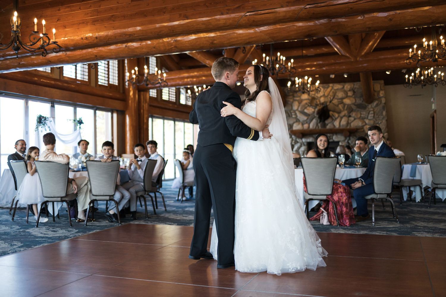 A romantic first dance at a rustic barn wedding reception with elegant chandeliers and wooden beams overhead.