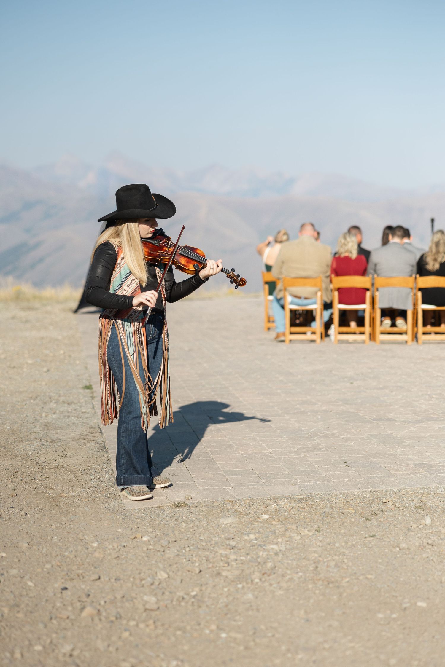 Musician playing violin outdoors with wooden chairs in background at sunset.