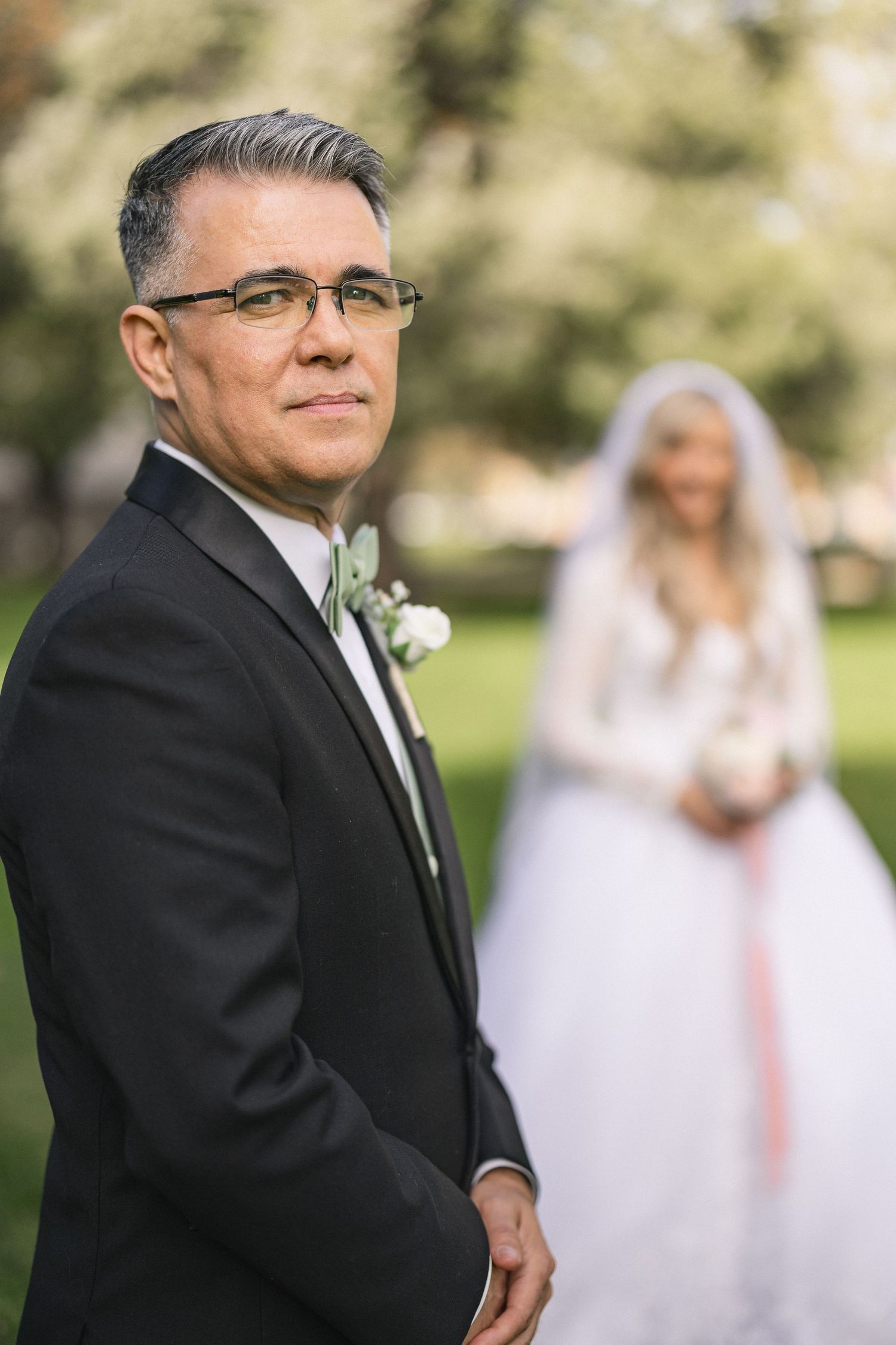 Groom in classic black tuxedo and glasses with bride in background at outdoor wedding.