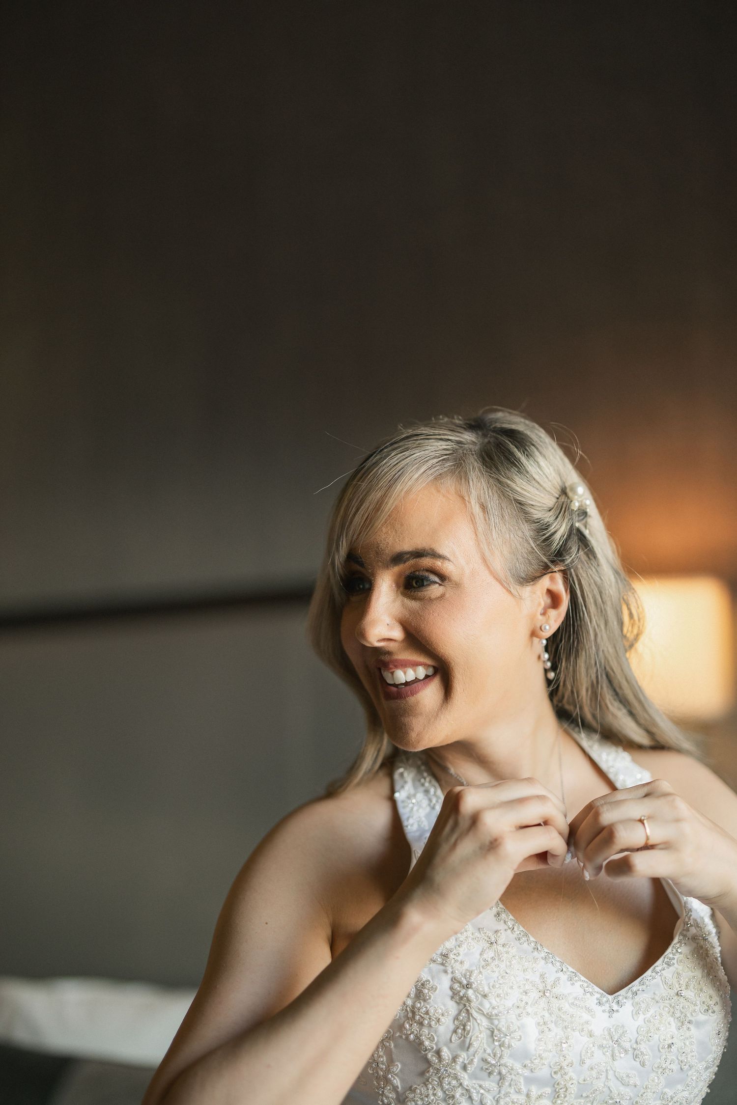 Joyful bride in white lace dress smiling while adjusting jewelry before wedding.