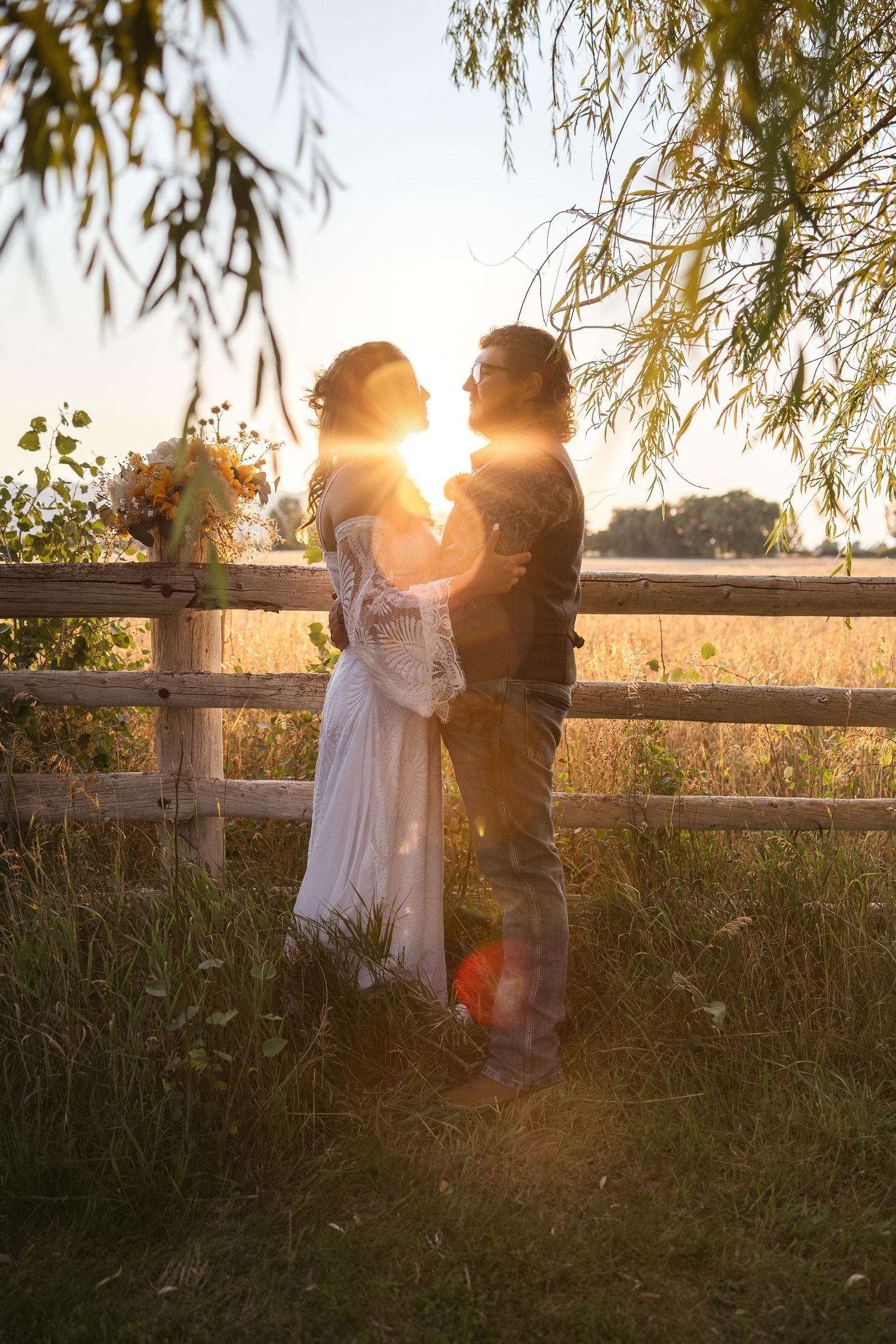 A couple shares a tender moment at sunset against a rustic fence with golden sunlight streaming through trees.