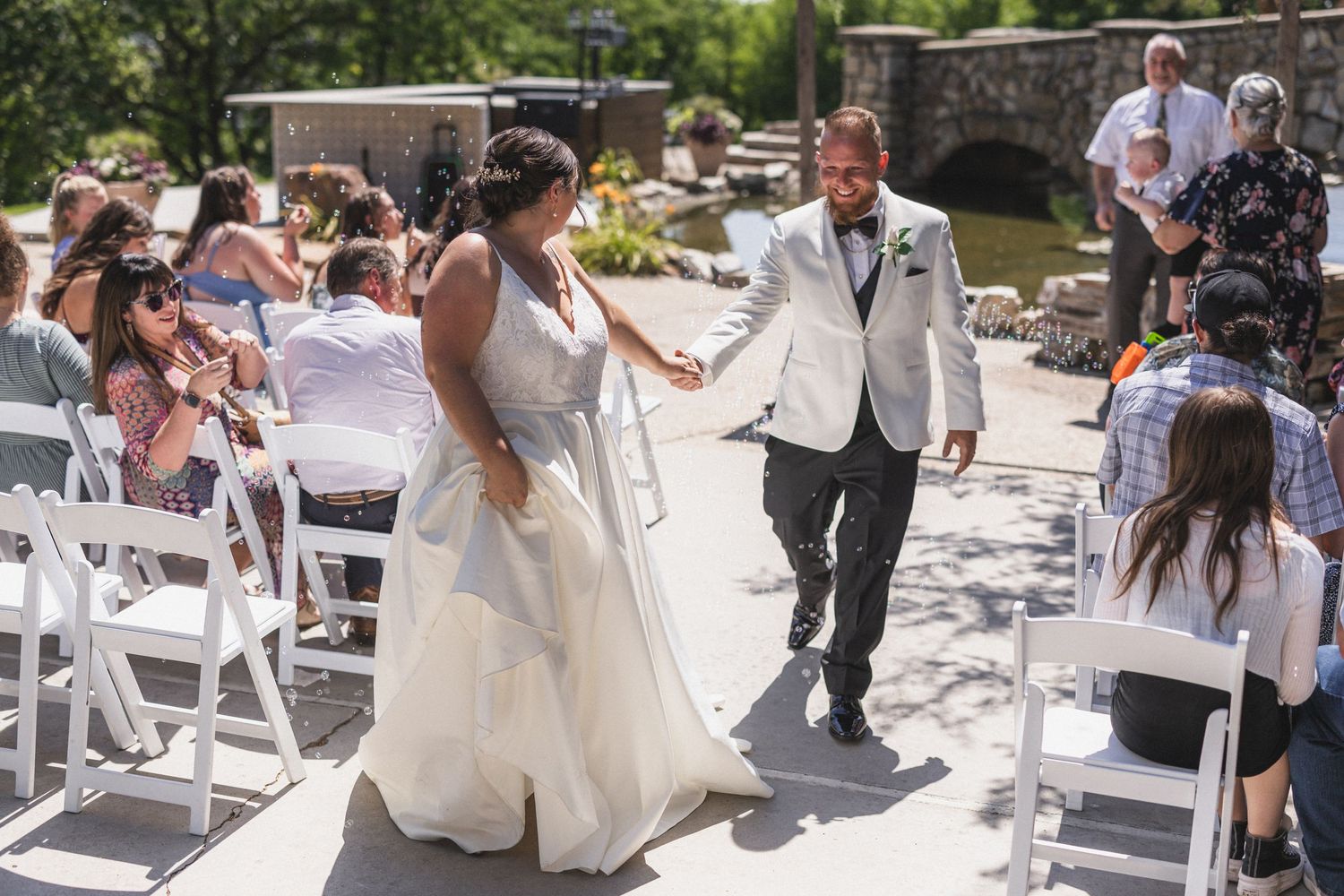Outdoor wedding ceremony with guests seated in white chairs on stone patio.