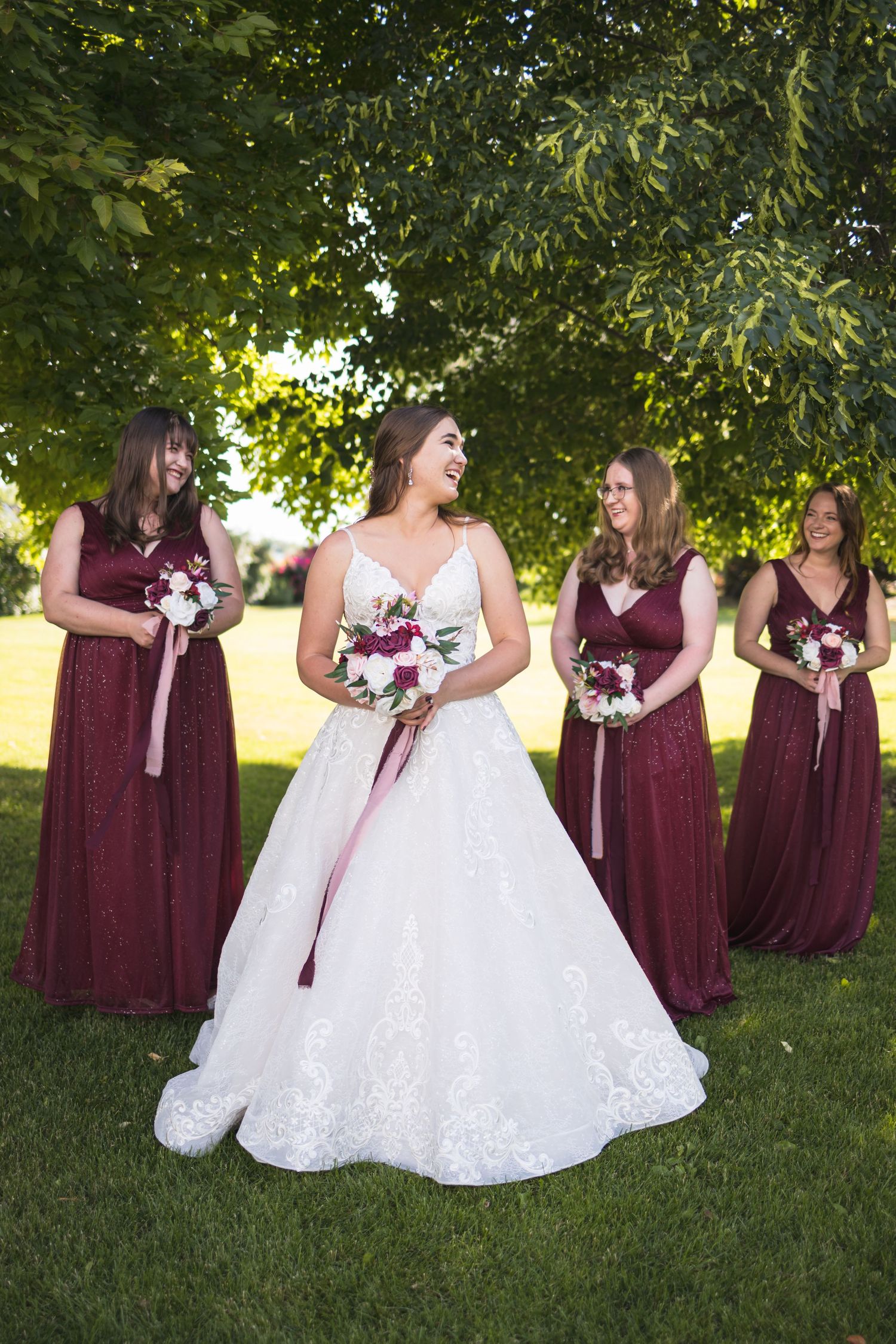 Wedding party portrait with bridesmaids in burgundy dresses holding bouquets on a sunny lawn.
