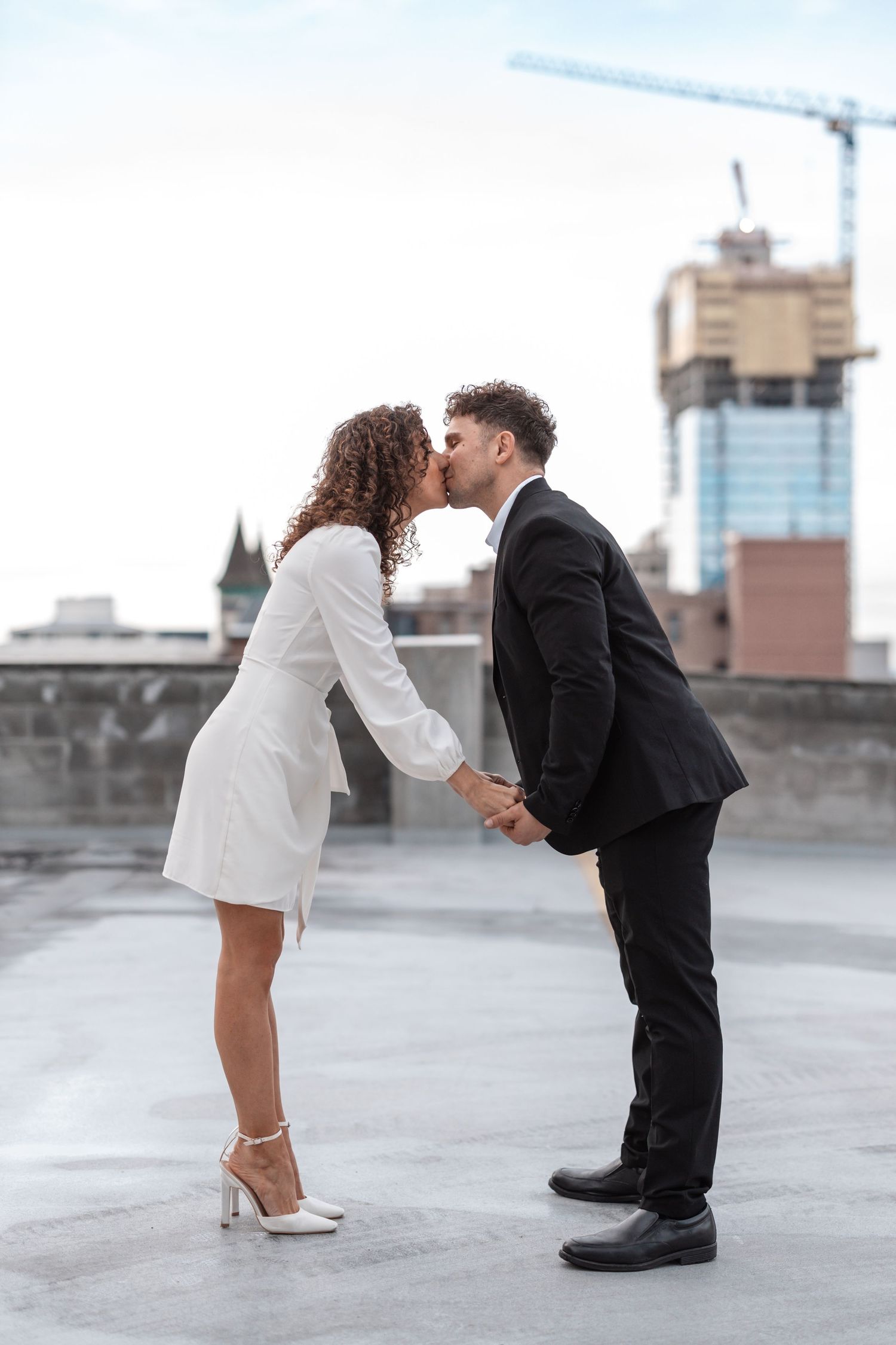 A couple in contrasting black and white outfits share a tender kiss on an urban rooftop with construction in background.