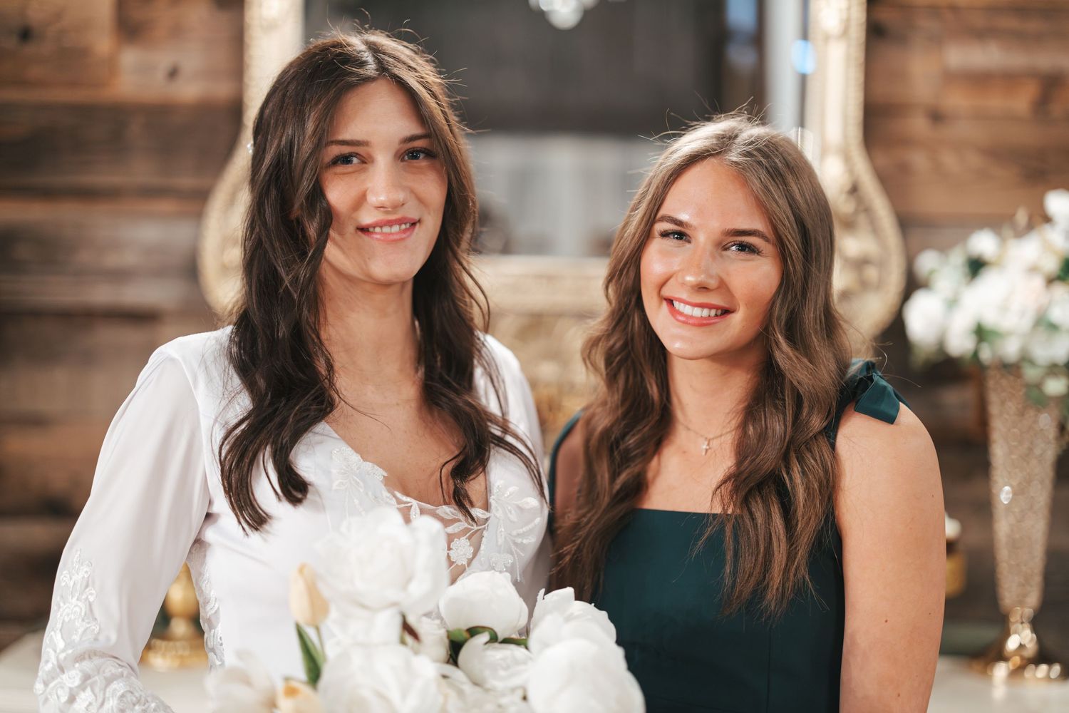 Two happy women in white and dark dresses pose with bridal bouquet.