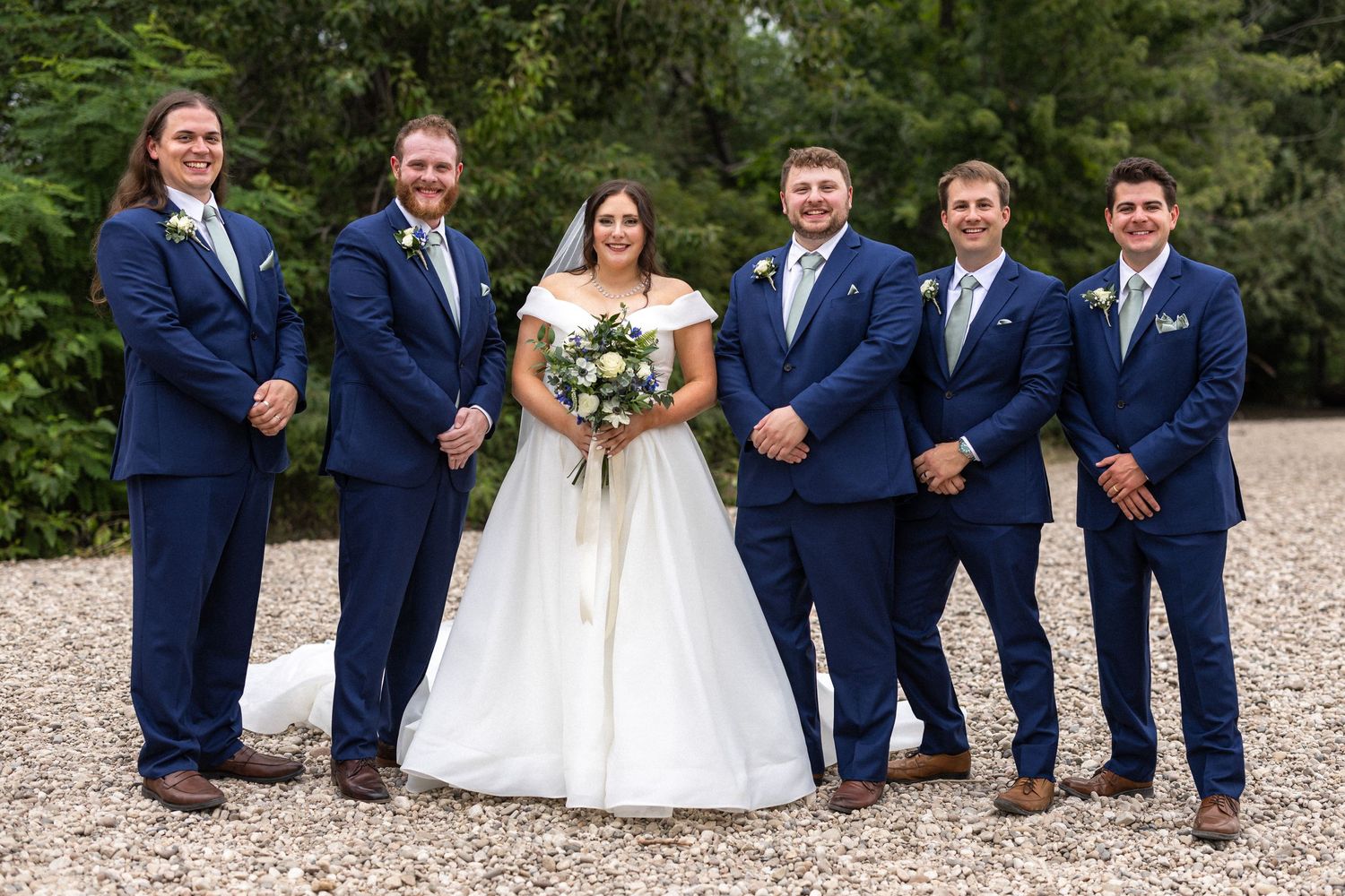 Wedding party in coordinated navy suits poses with bride in white gown on outdoor path.