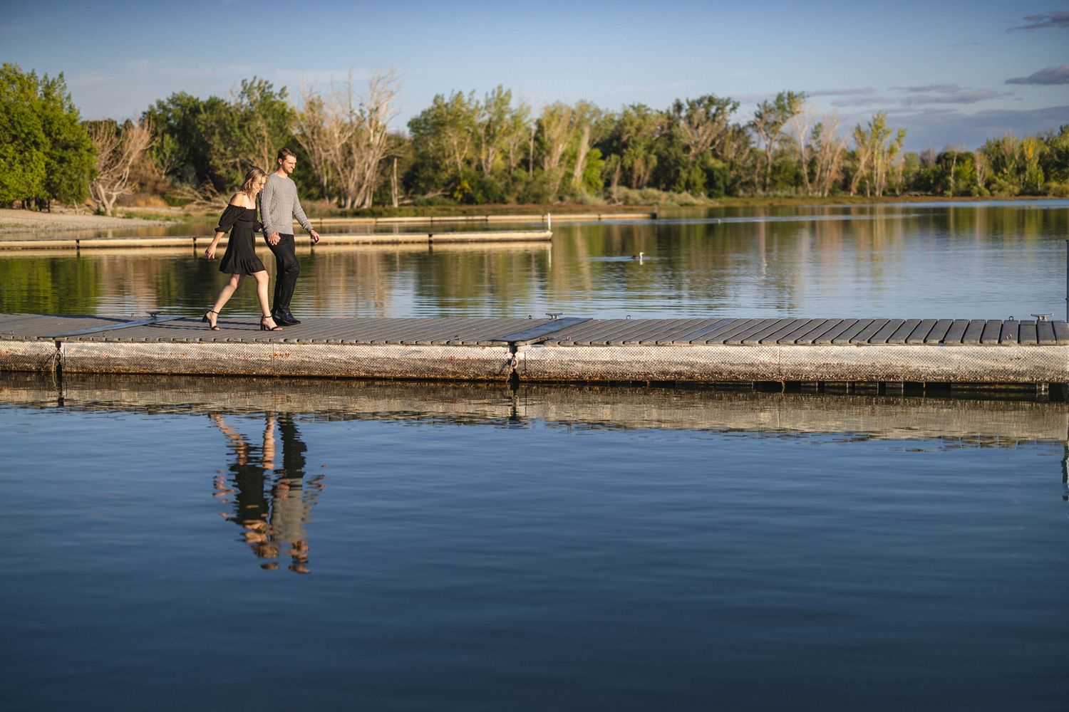 A couple sits on a wooden dock overlooking a peaceful lake surrounded by trees.