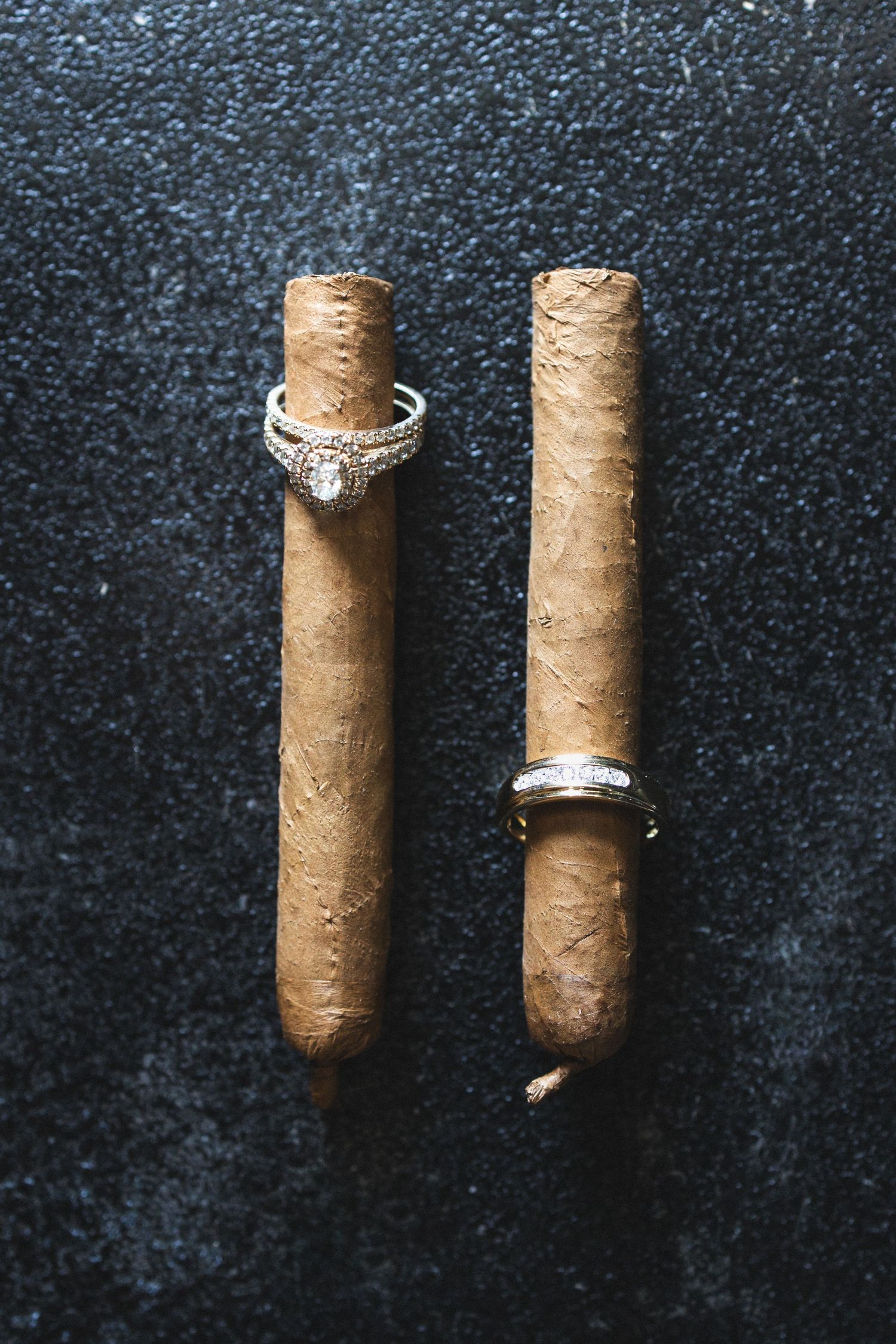 Wedding rings displayed creatively on two cigars against a dark textured background.