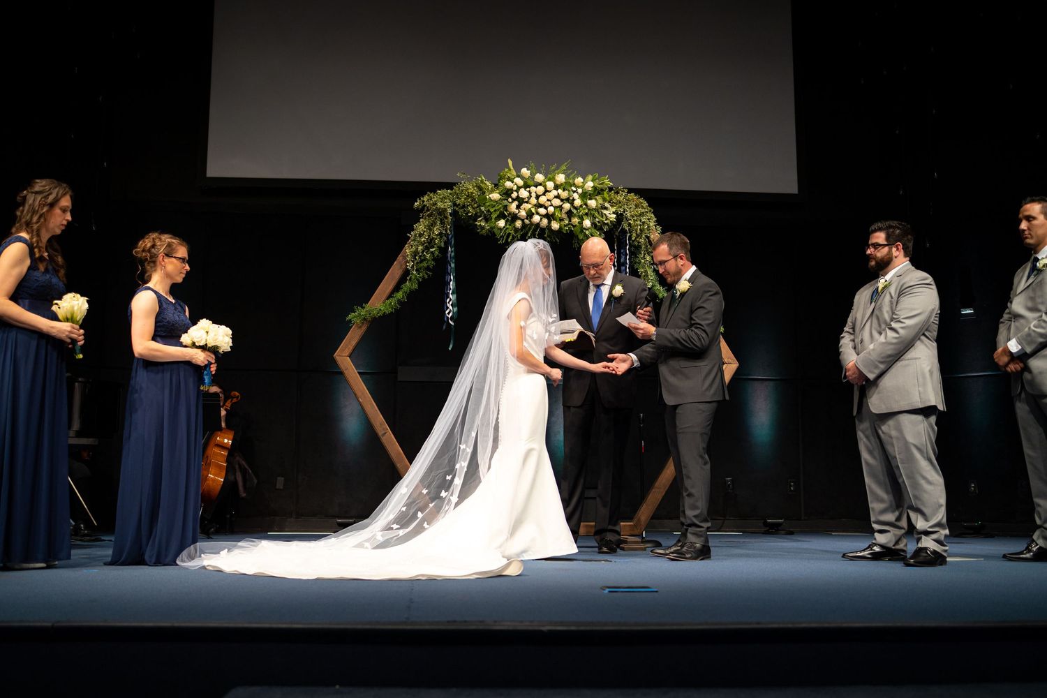 Wedding ceremony taking place under a hexagonal floral arch with wedding party in navy and gray attire.