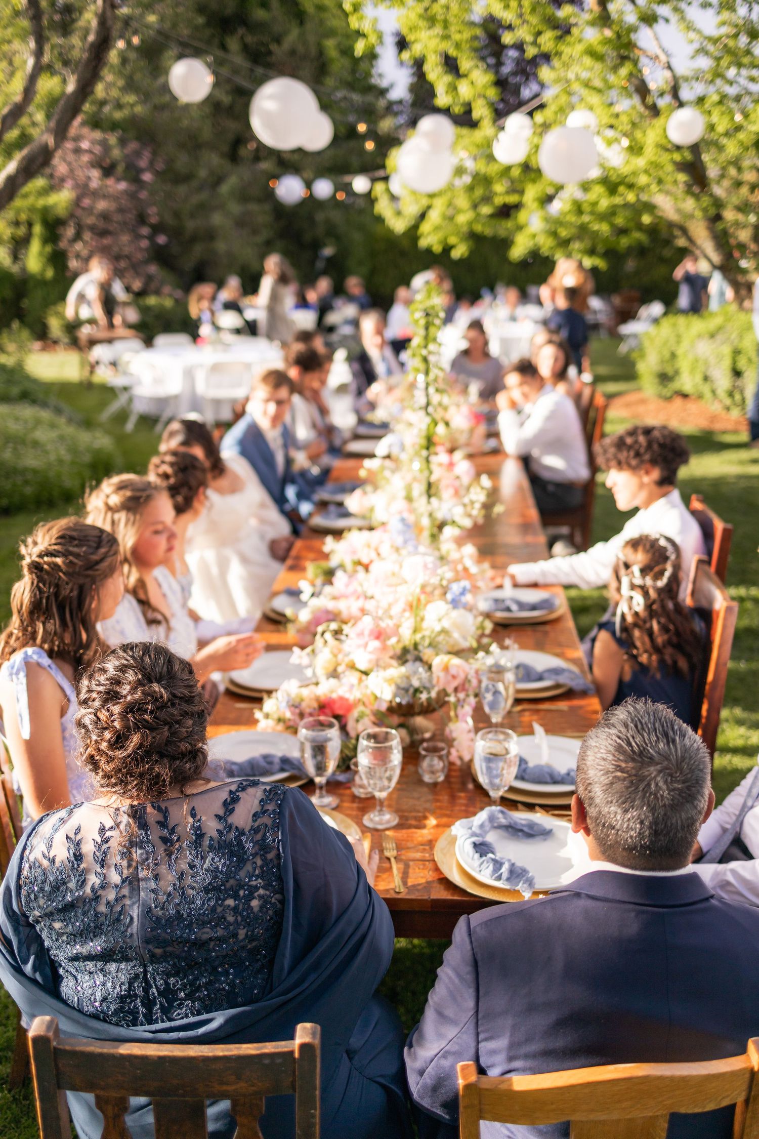 Outdoor wedding reception features long wooden table with string lights and floral centerpieces.