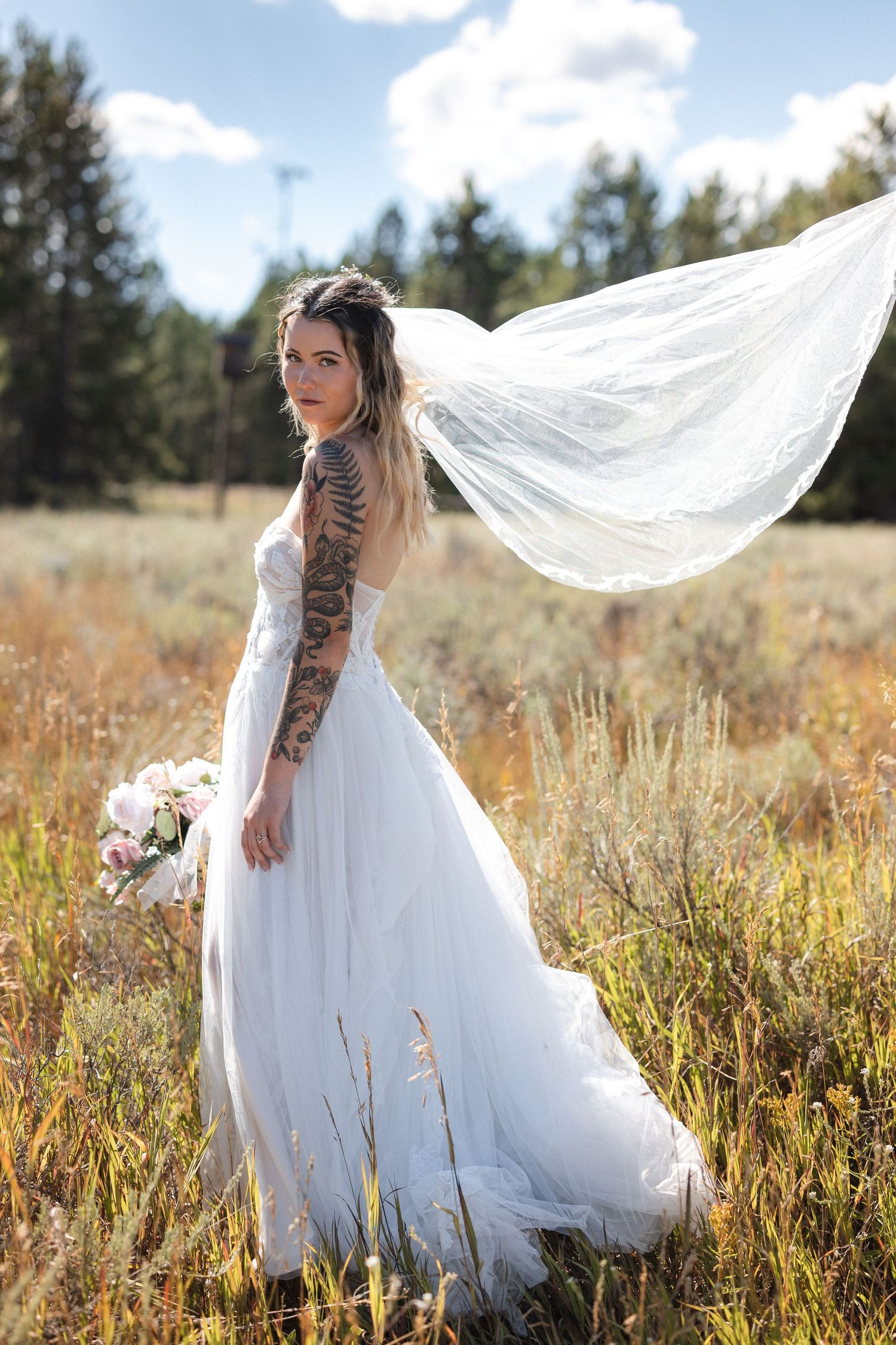 A bride in a flowing white dress poses in a field with her veil floating in the breeze.