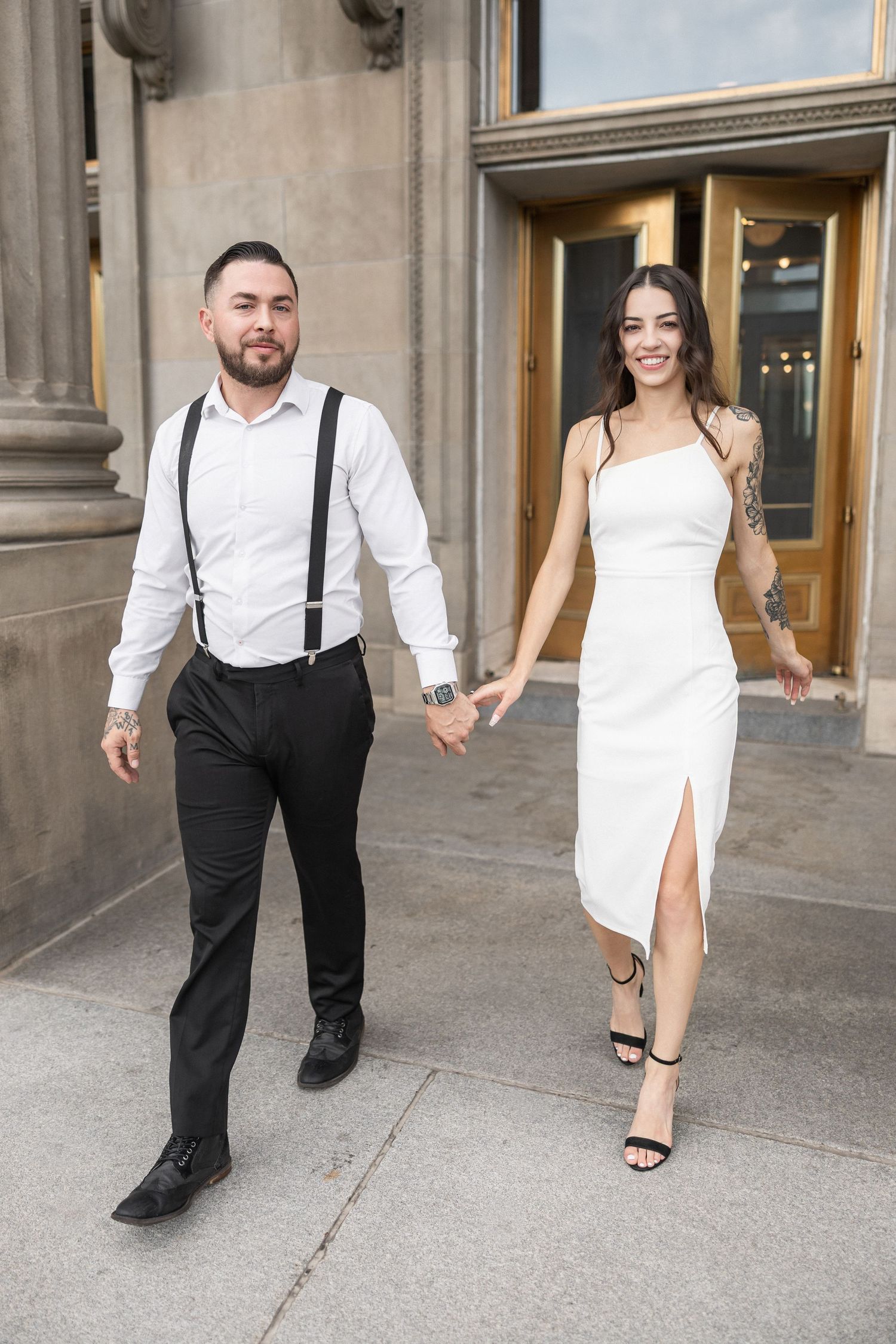 A couple in formal attire walks together outside a classical building entrance.