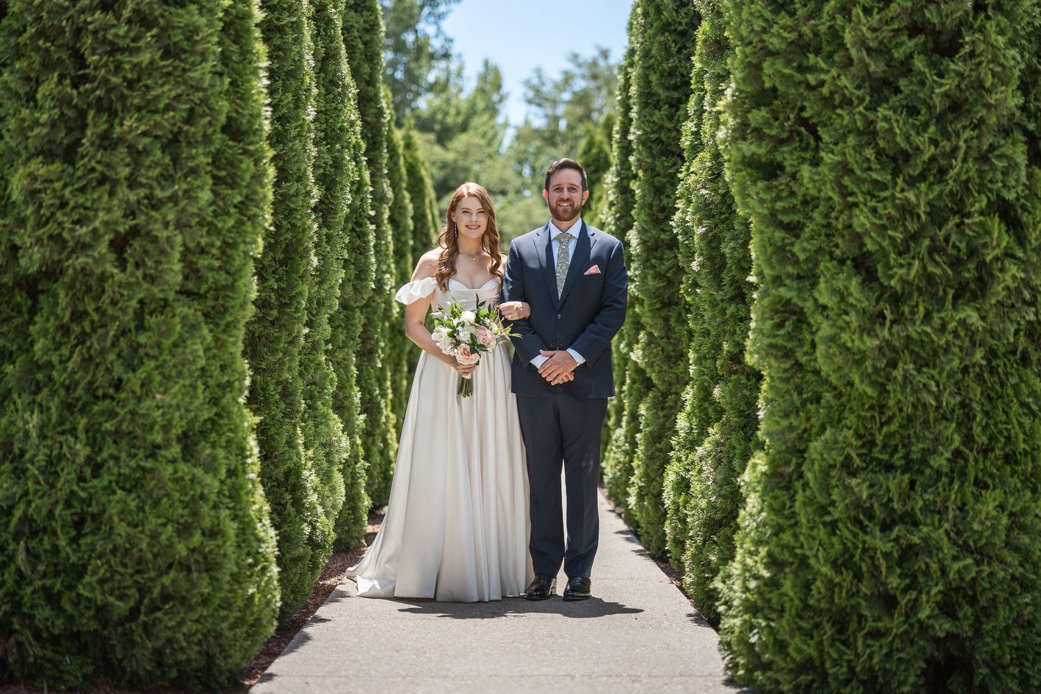 A couple poses together on a walkway lined with towering green cypress trees.