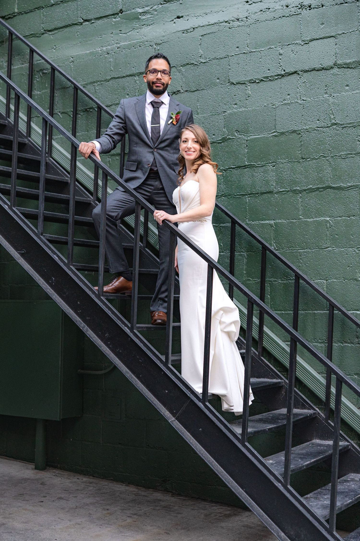 A wedding couple poses on an industrial metal staircase against a green wall.