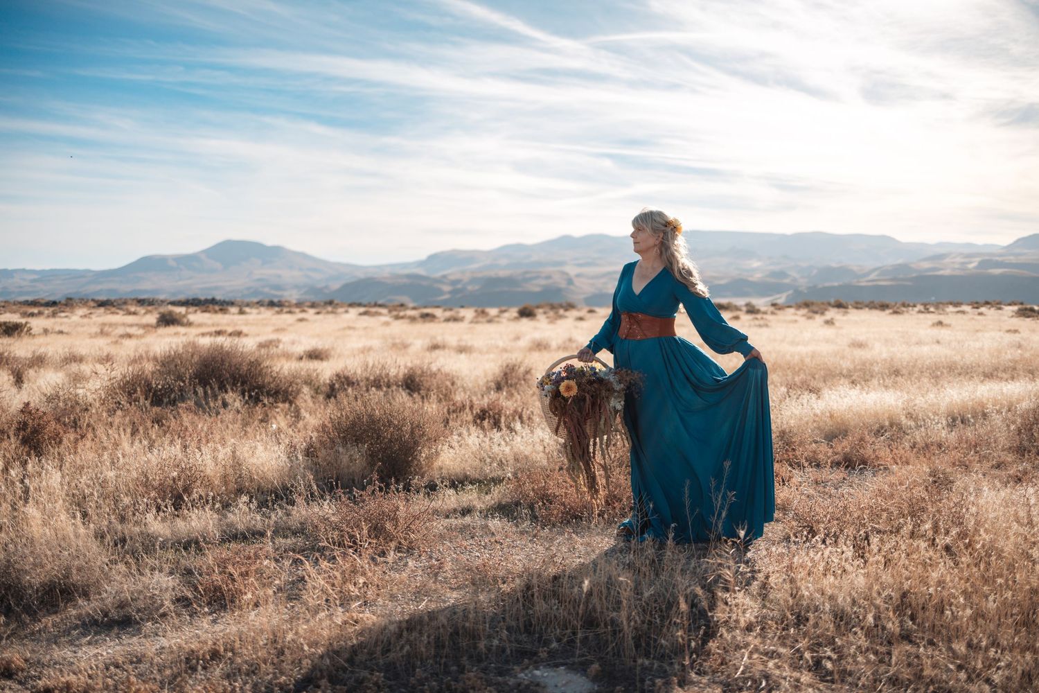 A figure in a teal dress sits gracefully in a golden field with mountains and dramatic sky in the background.