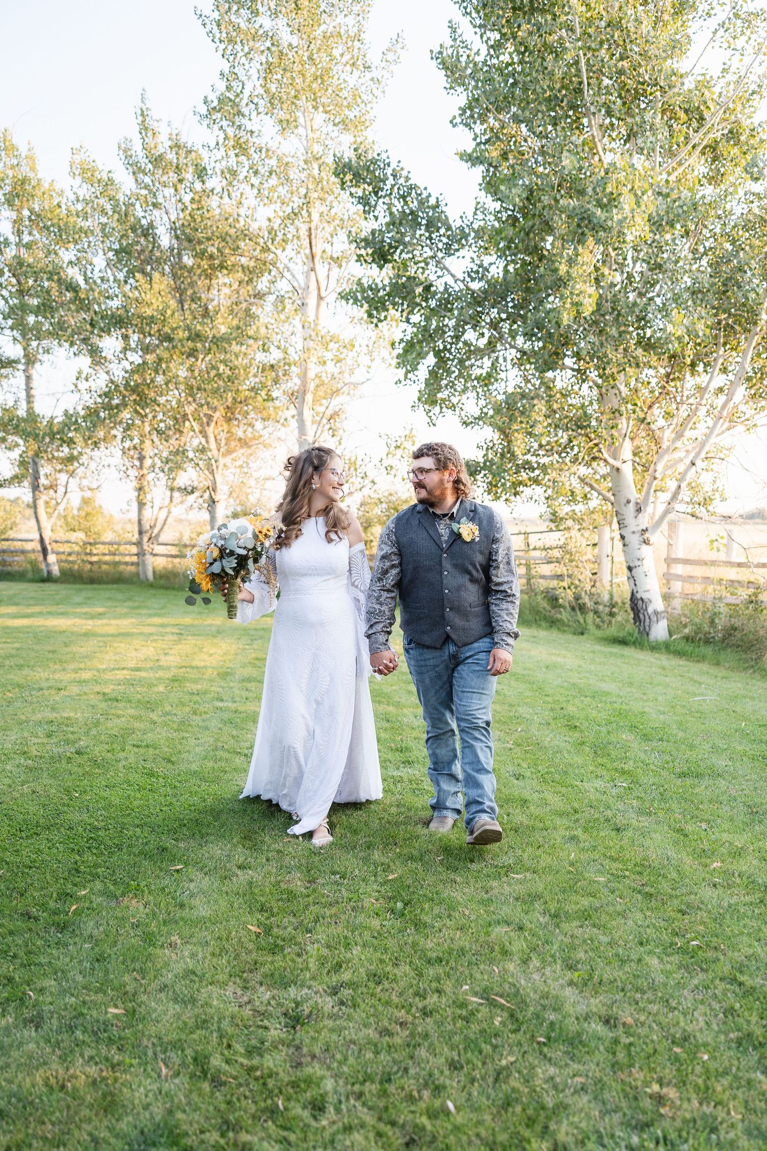A couple walks hand in hand on green grass with white birch trees in the background during a rustic outdoor wedding.