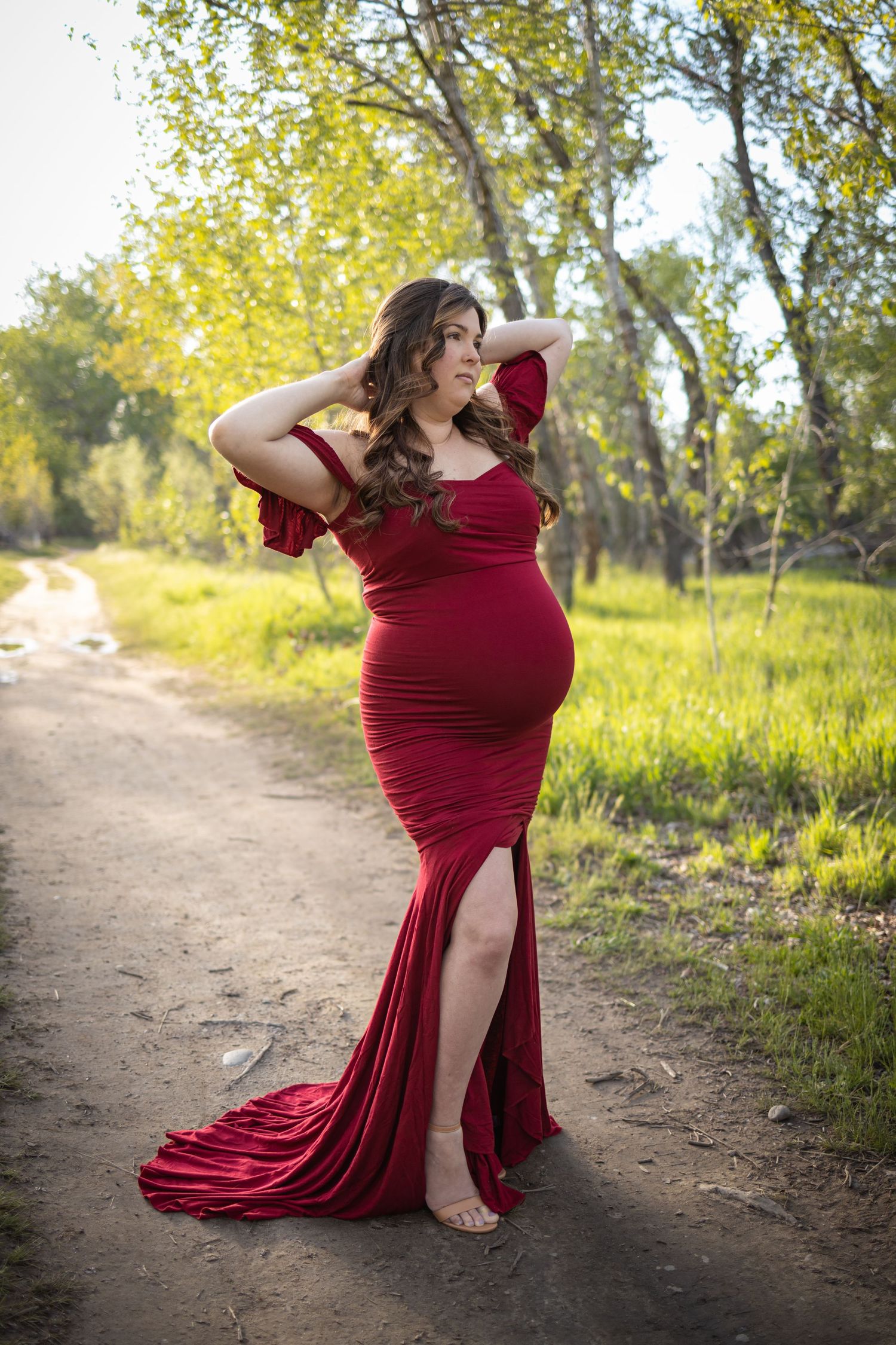 A figure in a flowing burgundy gown poses gracefully on a dirt path surrounded by sunlit trees and greenery.
