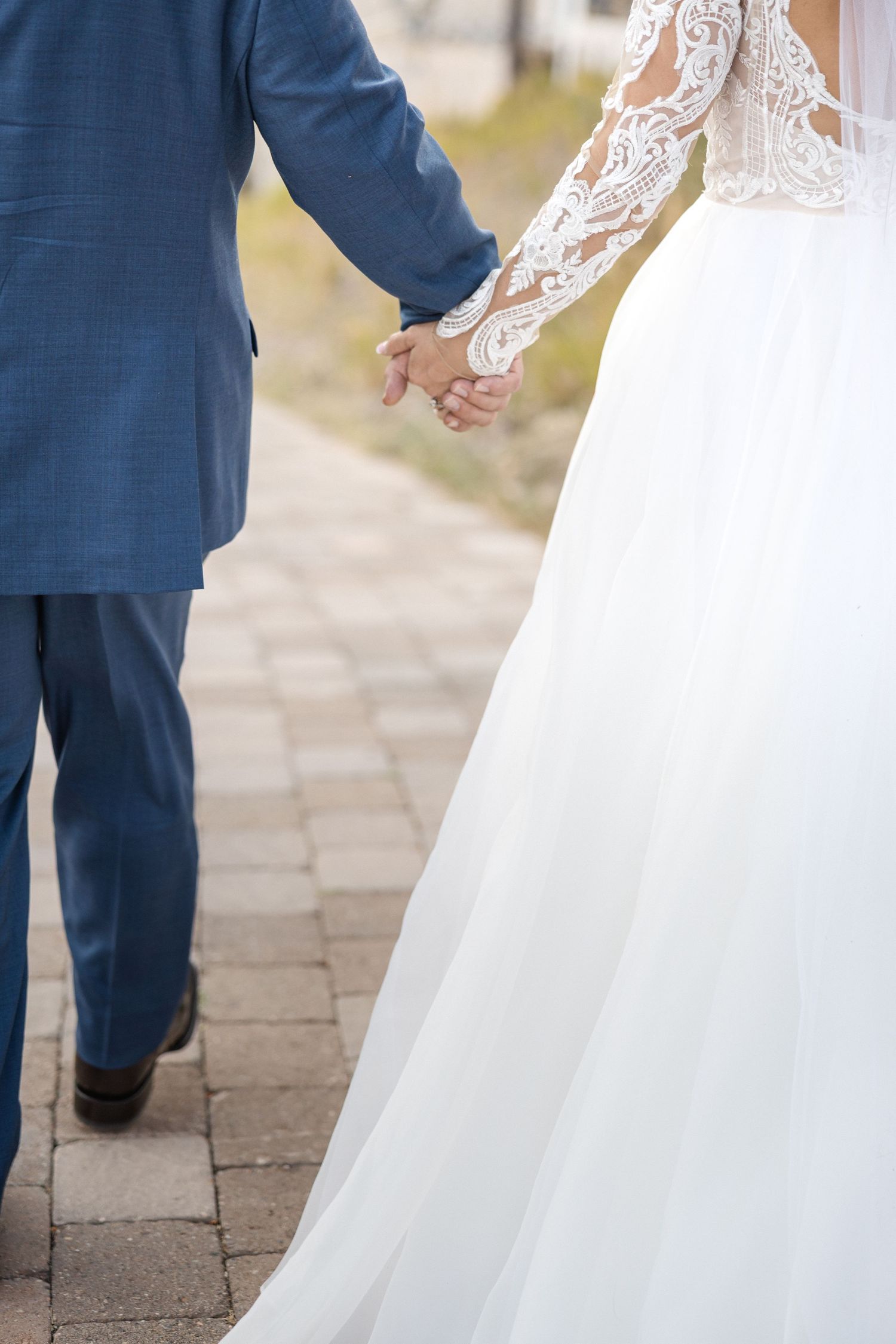 Close-up details of a bride's lace wedding dress and groom's navy suit as they hold hands.