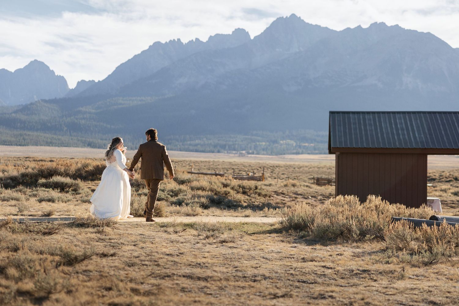Newlyweds walk through a rustic mountain landscape with dramatic peaks in the background.