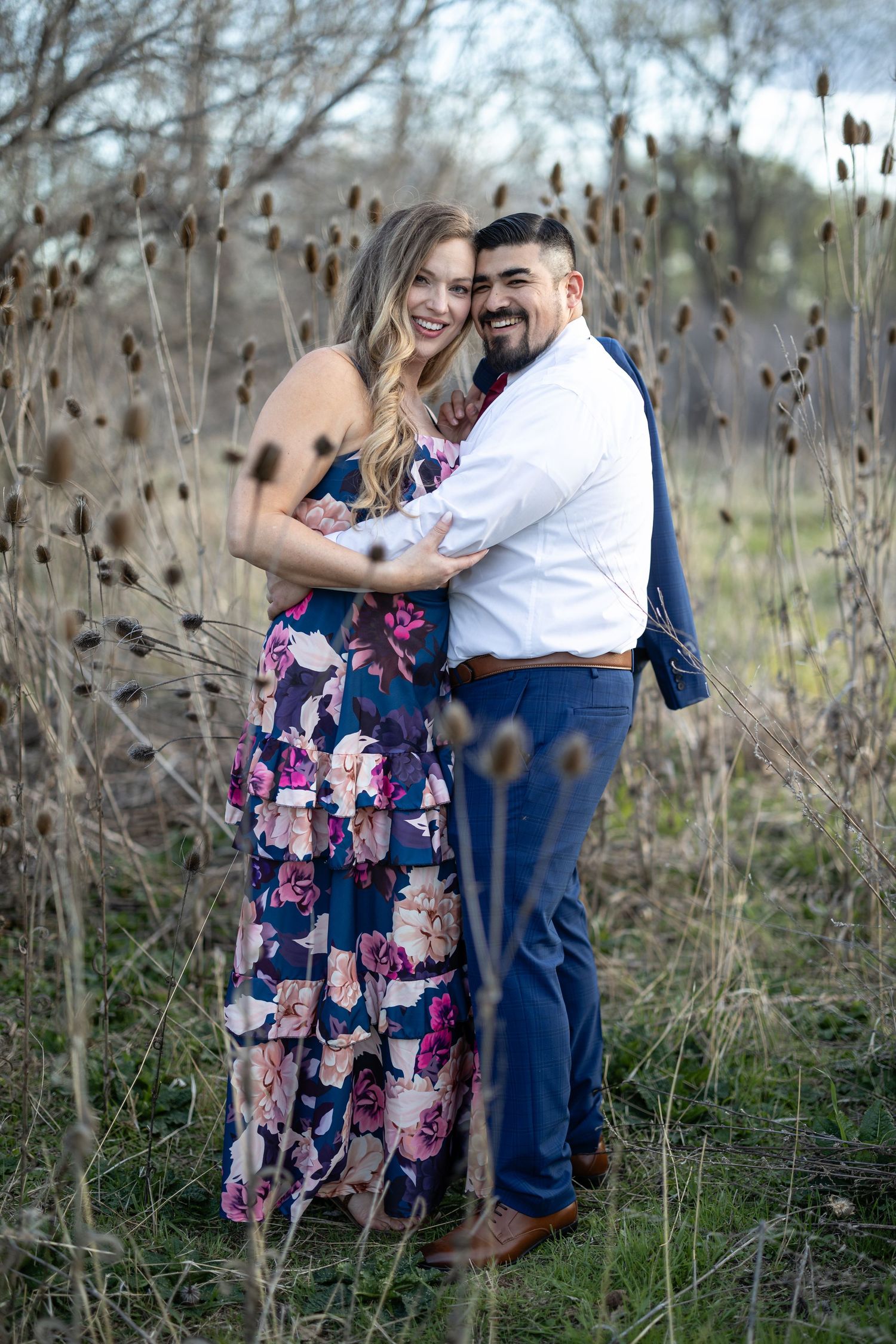 Happy couple embraces while standing among dried grass and wildflowers in a natural setting.