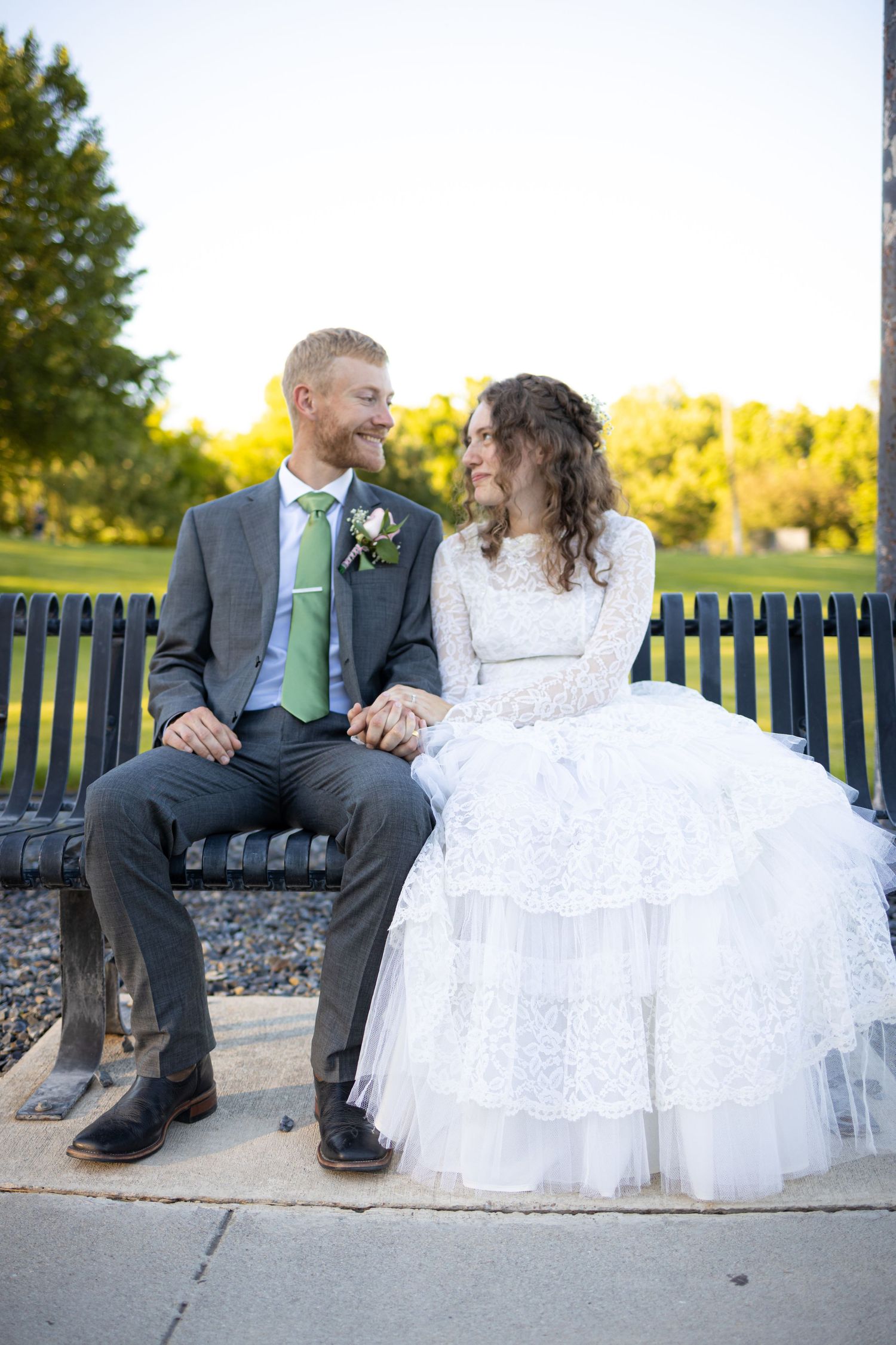 Couple sitting on park bench in wedding attire sharing intimate moment at sunset.