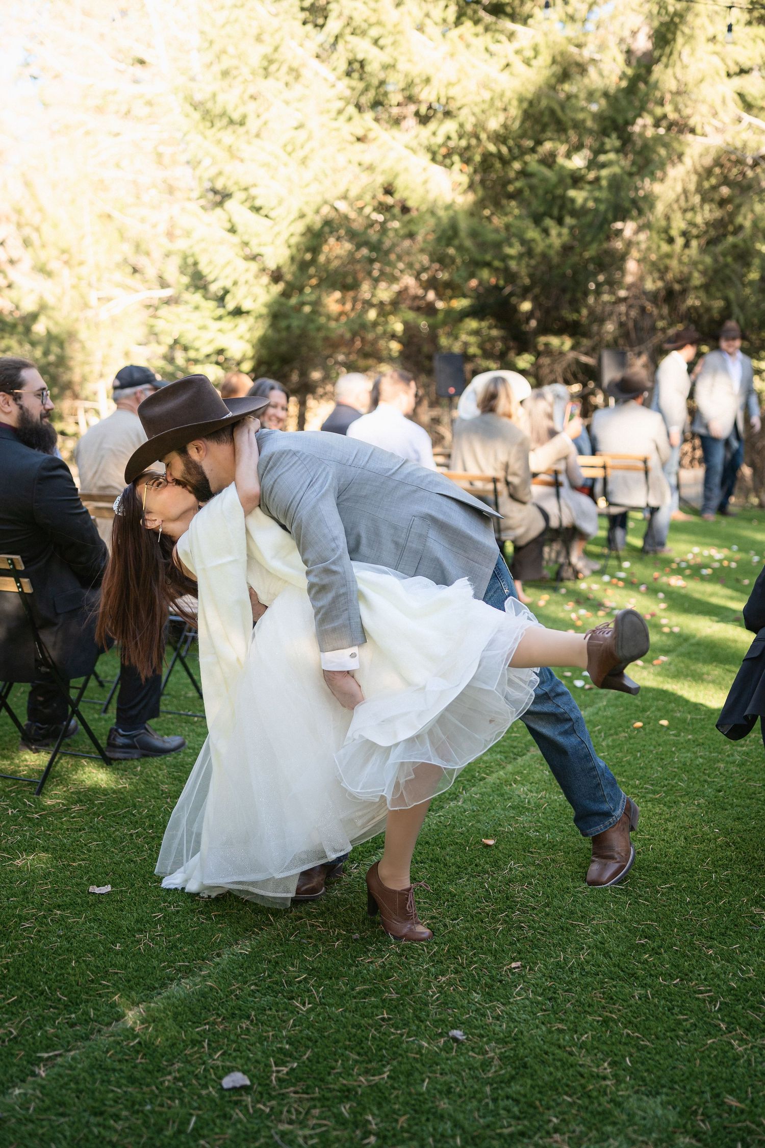 A couple performs a romantic dip pose during an outdoor wedding reception on a grassy lawn with guests seated nearby.