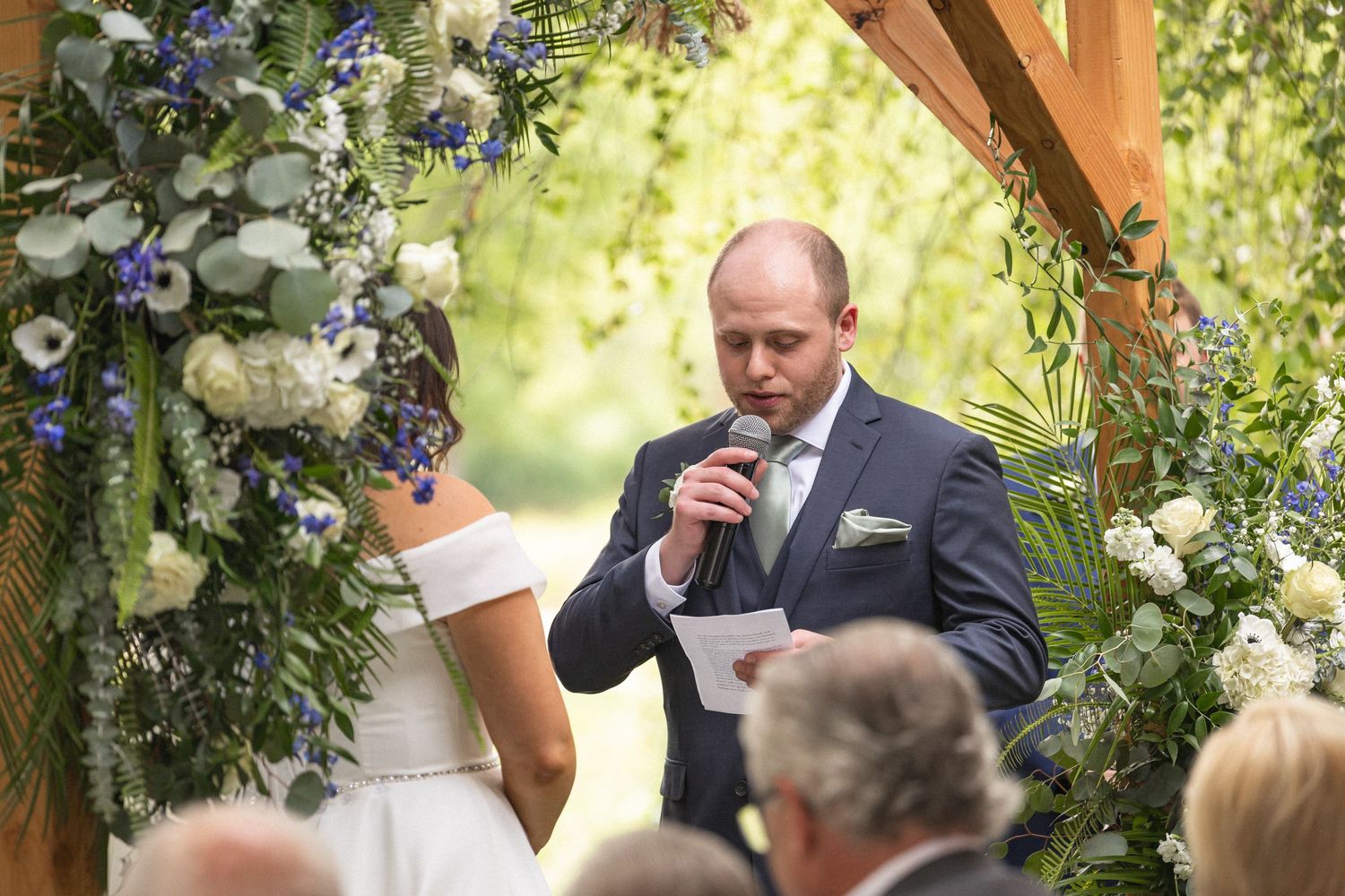 Wedding ceremony under floral-decorated wooden arch with guests seated outdoors.