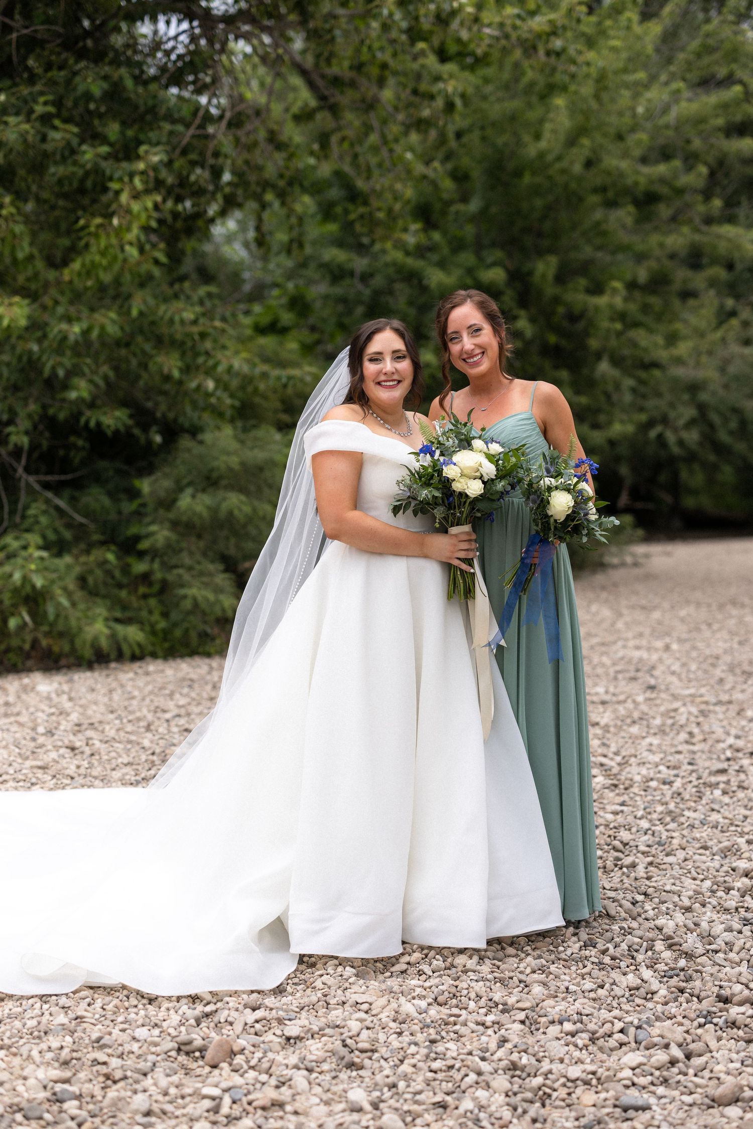 Bridesmaids in white and sage green dresses share joyful moments on gravel path surrounded by greenery.
