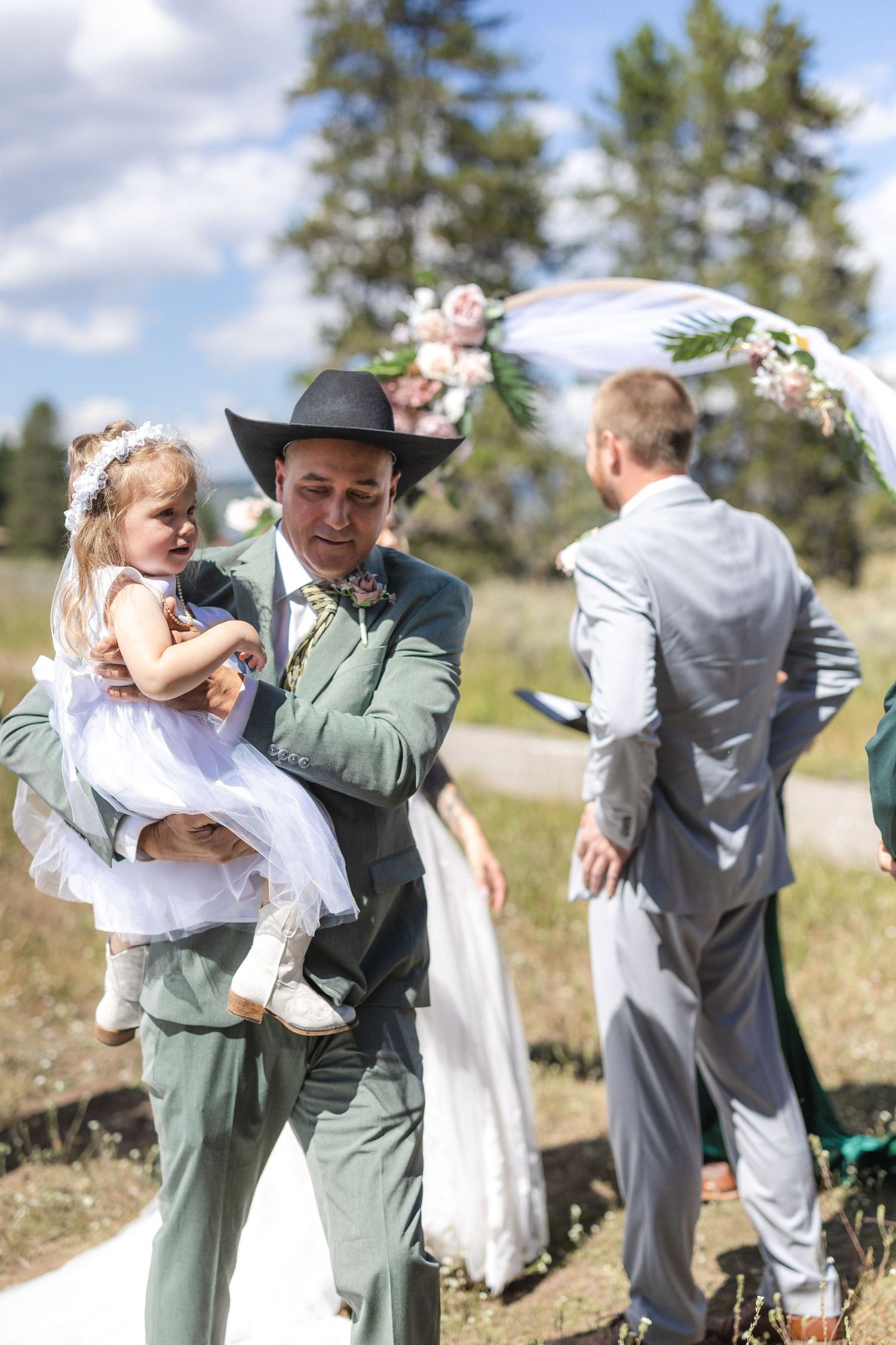 Wedding party gathered outdoors with children in formal attire beneath evergreen trees.