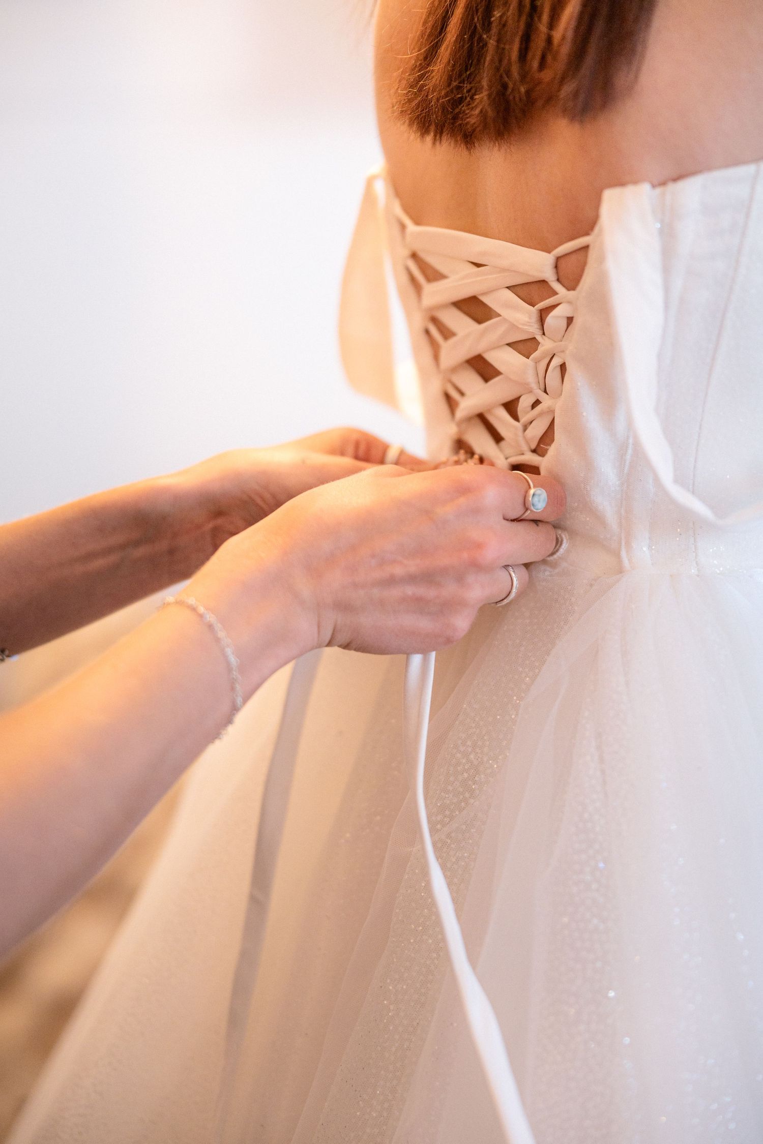 Hands lacing up the corset back of a white wedding dress in soft lighting.