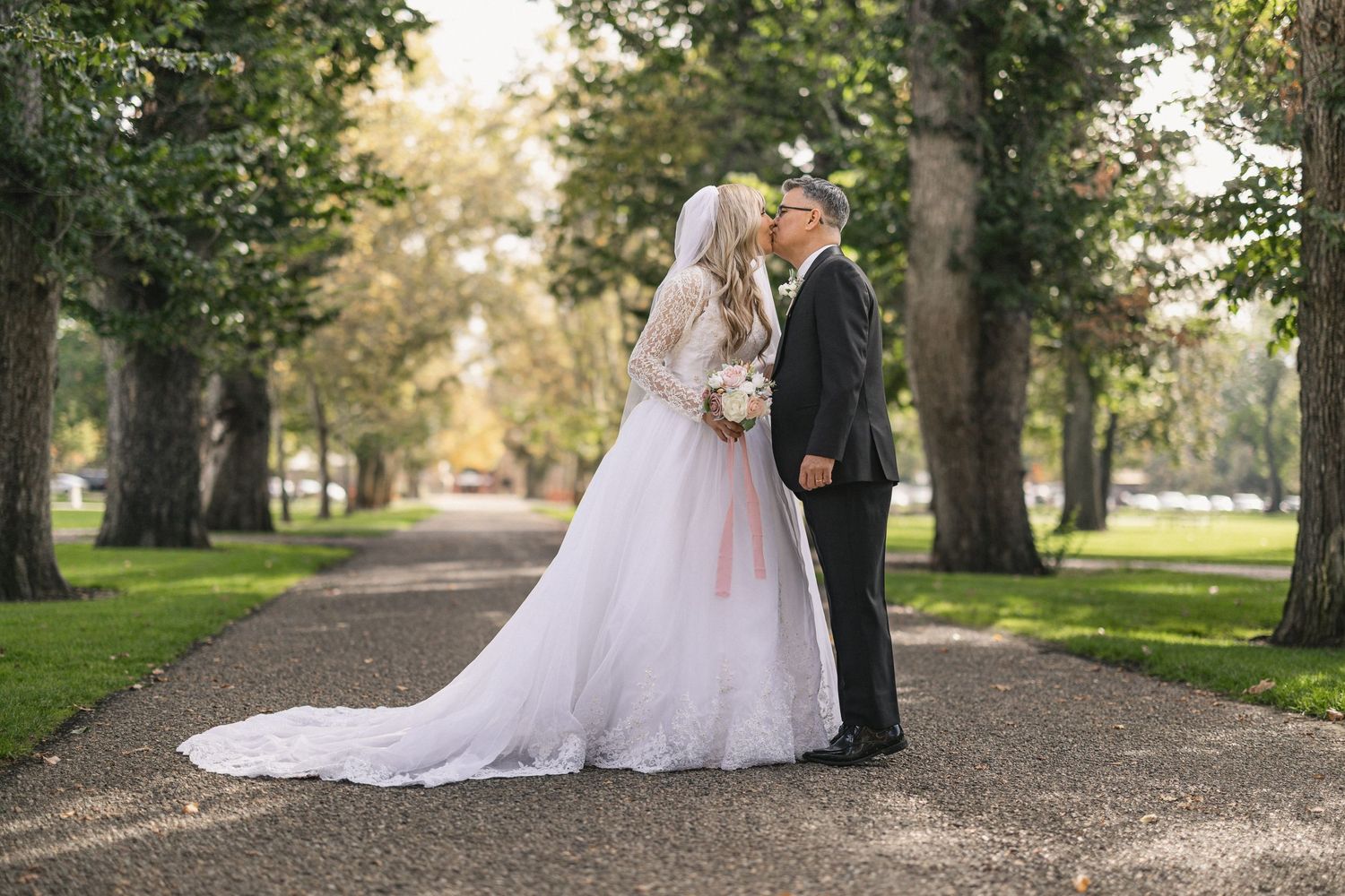 Romantic wedding portrait on a tree-lined path with soft natural lighting.