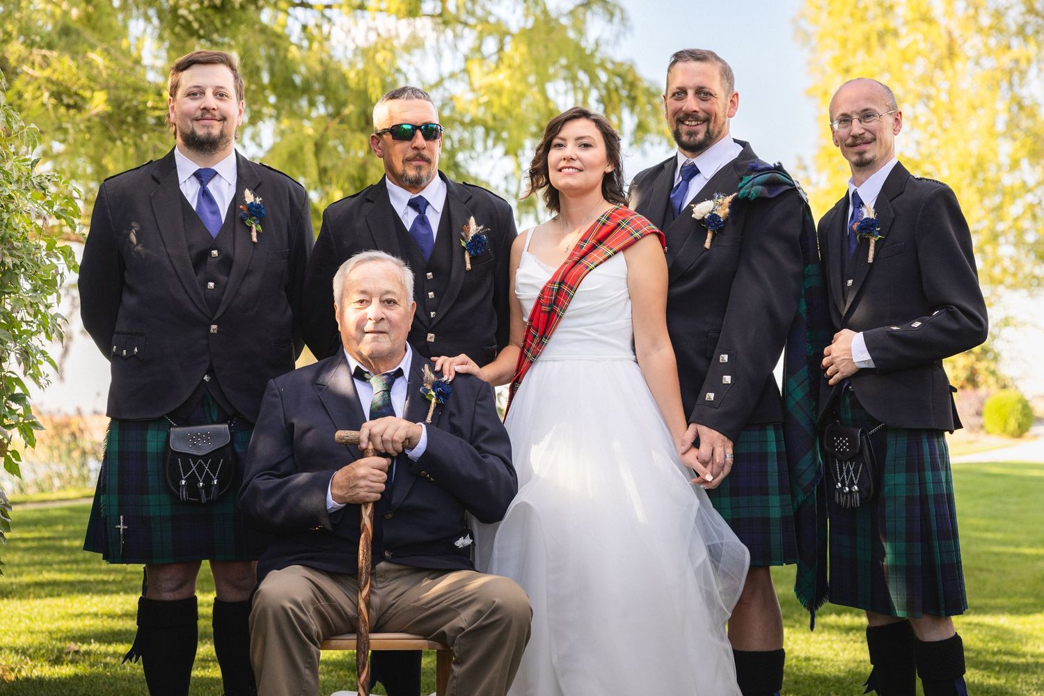 Wedding party poses together outdoors with kilts and formal attire against autumn foliage.
