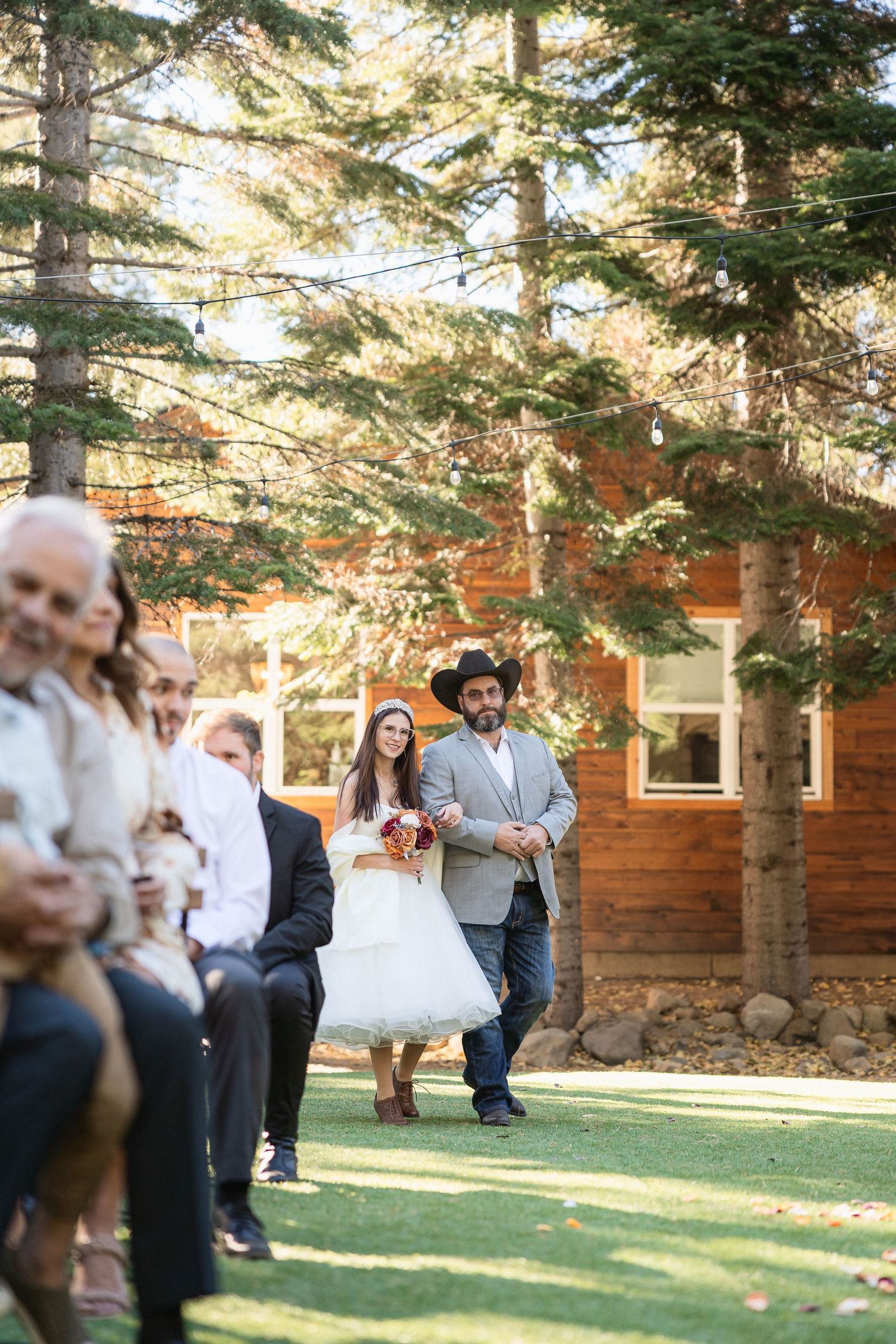 Guests enjoying outdoor wedding festivities near rustic log cabin with string lights.