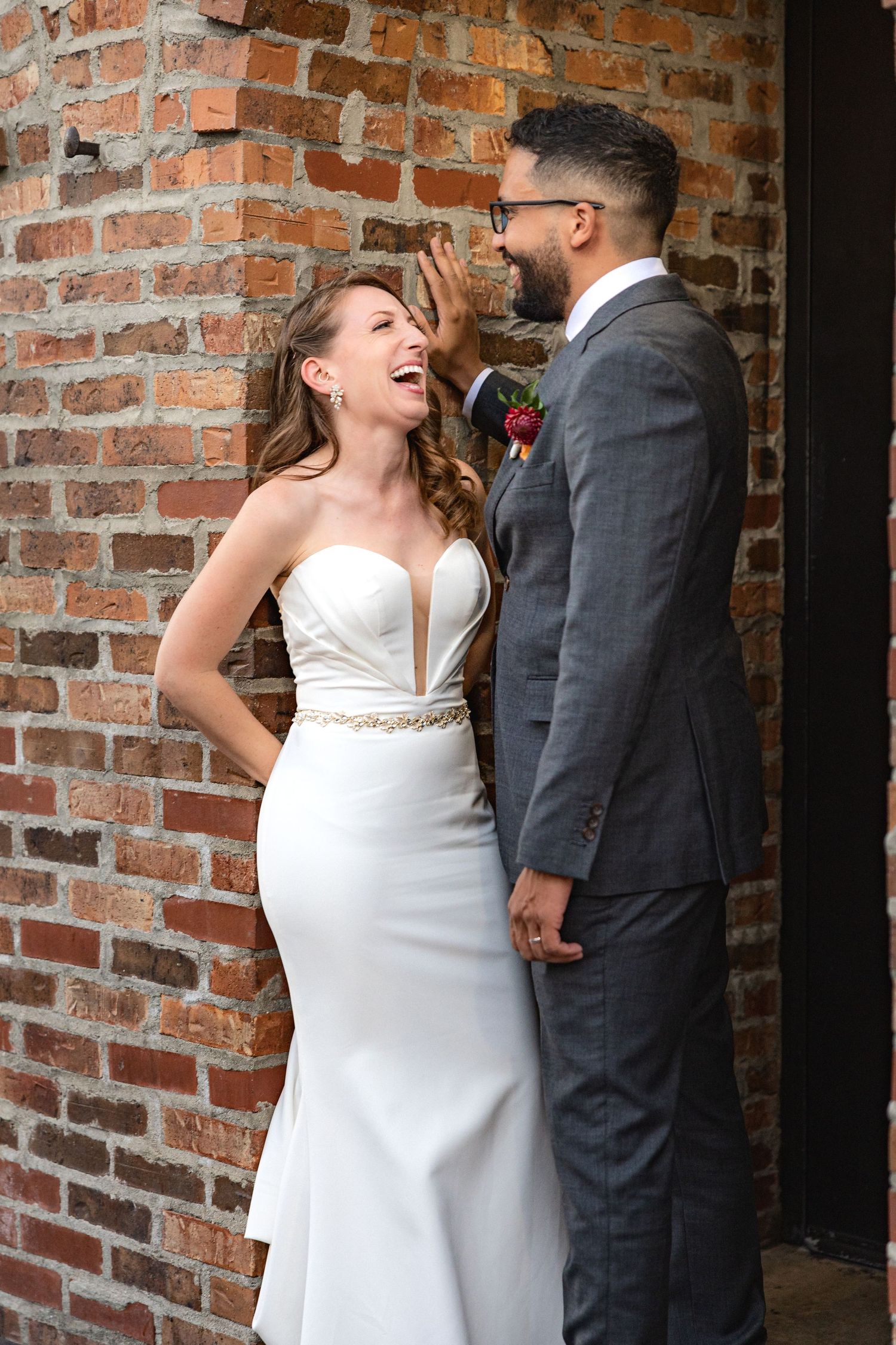 Wedding couple laughs together against rustic brick wall background.