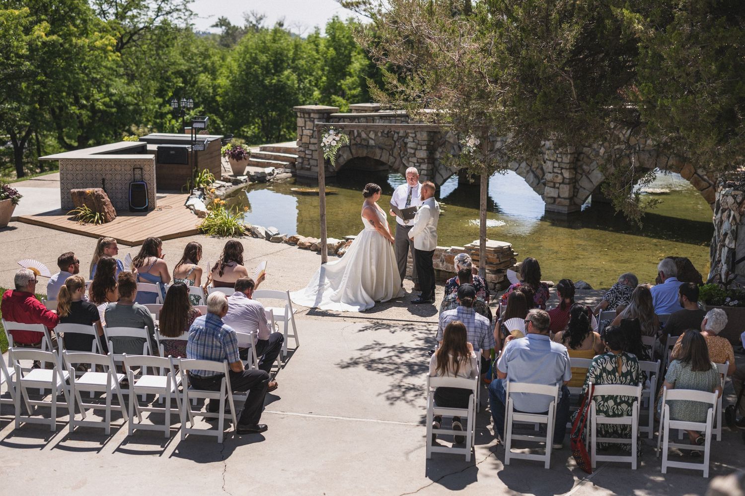 An outdoor wedding ceremony with guests seated in white chairs facing the couple and officiant.