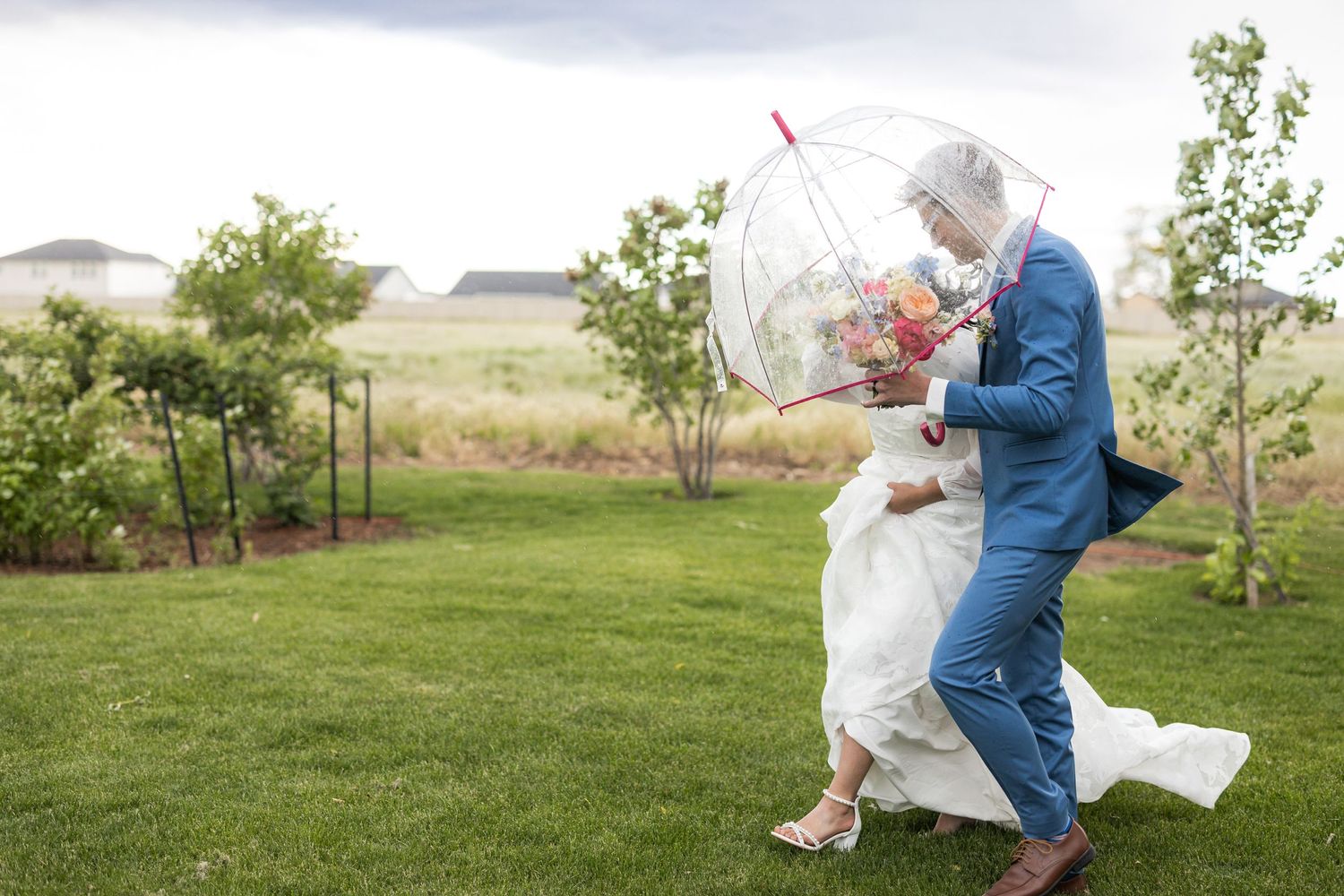 A windy wedding photo moment captured with couple sharing clear umbrella in outdoor garden setting.