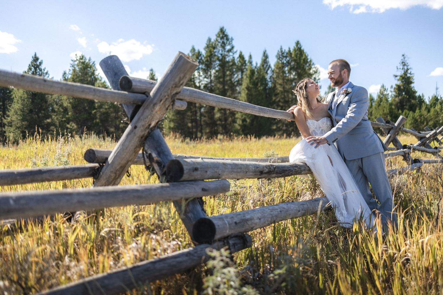 Playful wedding portraits taken along a wooden fence in a scenic mountain meadow.
