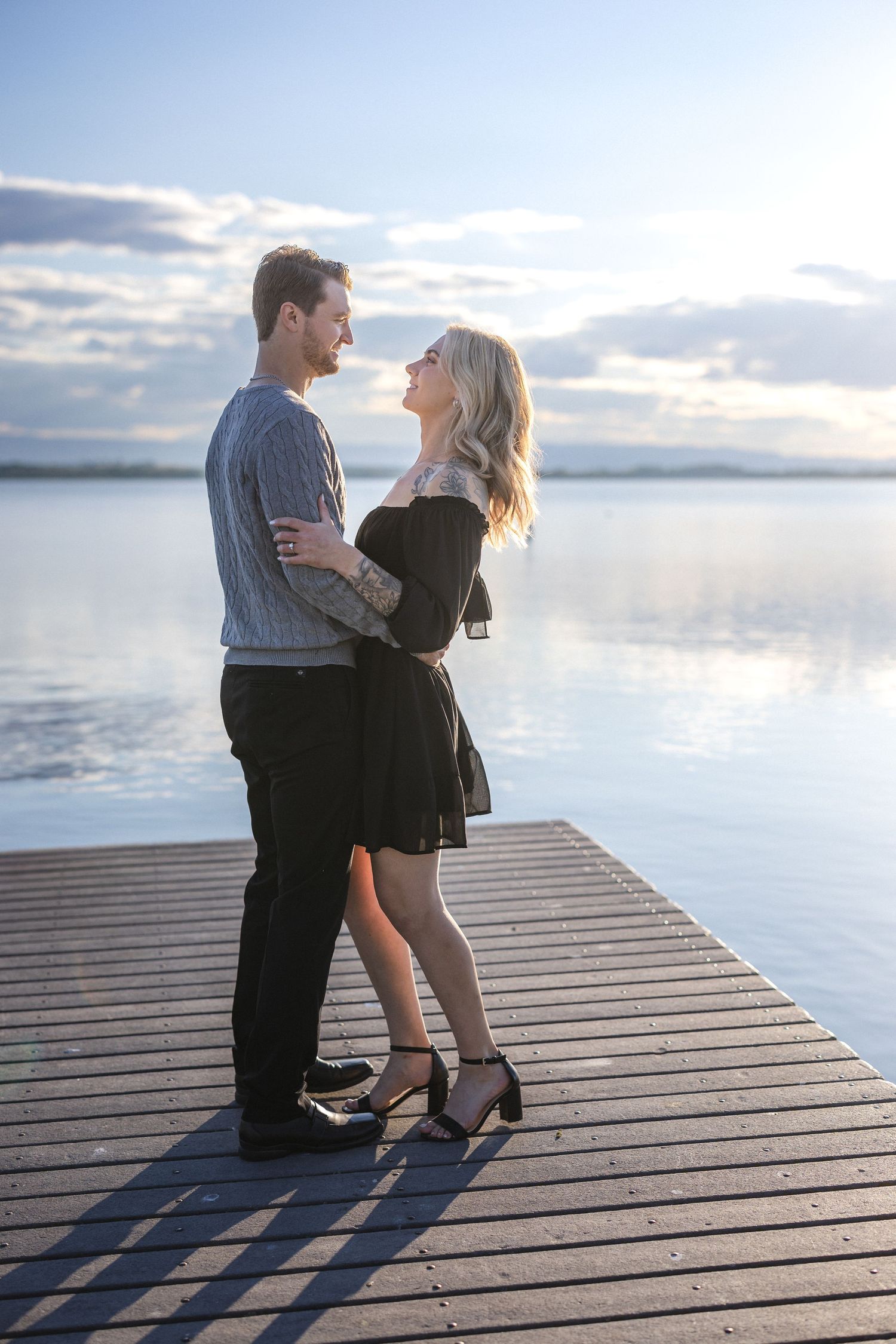 Couple embracing on wooden dock with scenic lake and cloudy sky backdrop.