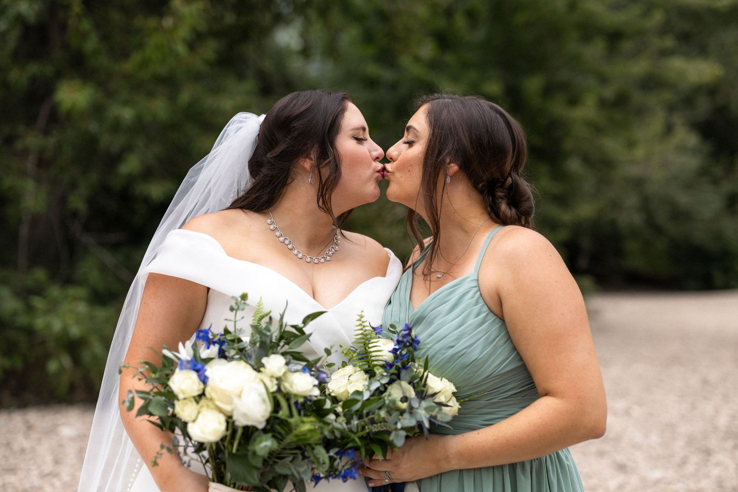Wedding party members in white and sage green dresses embrace while holding floral bouquets outdoors.
