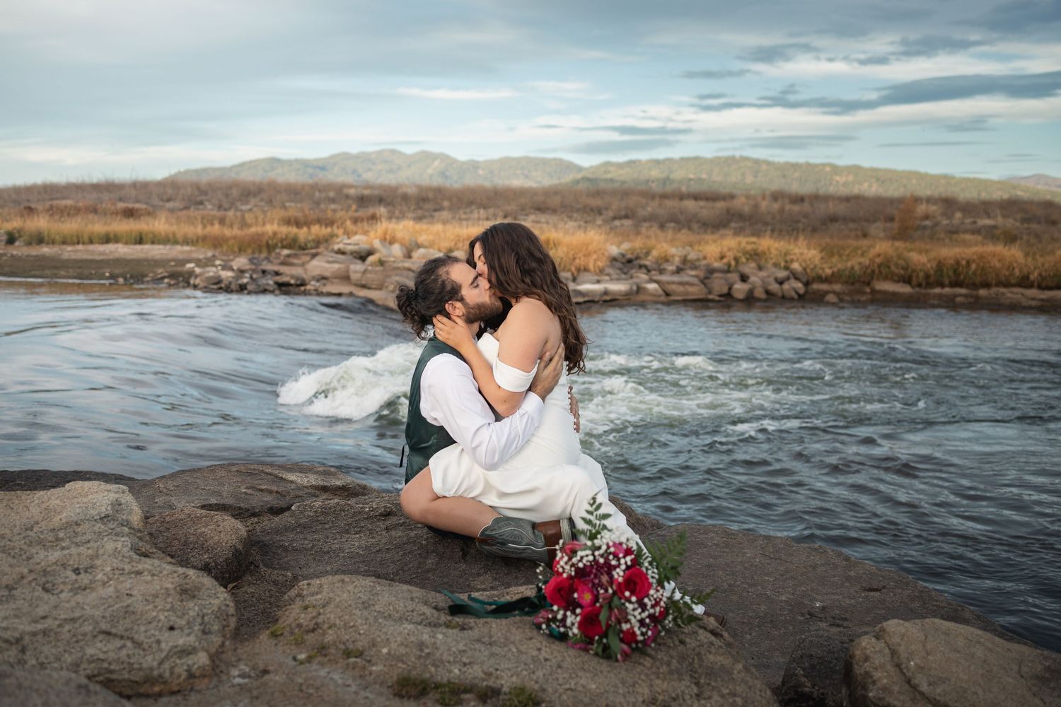 Intimate wedding moment captured on rocky riverside with mountains and flowing water in background.