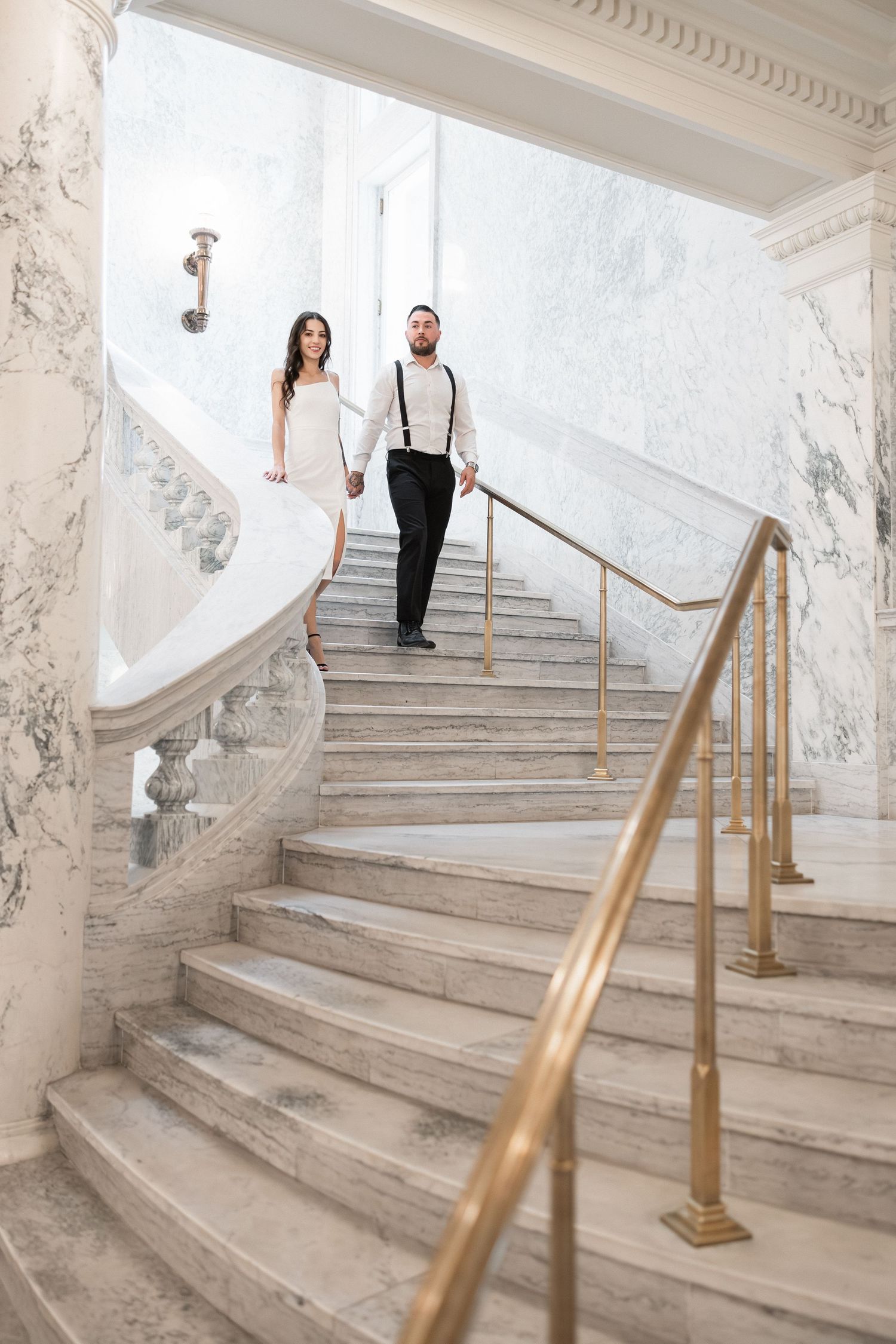 A couple descends a grand marble staircase in formal attire at an elegant venue.