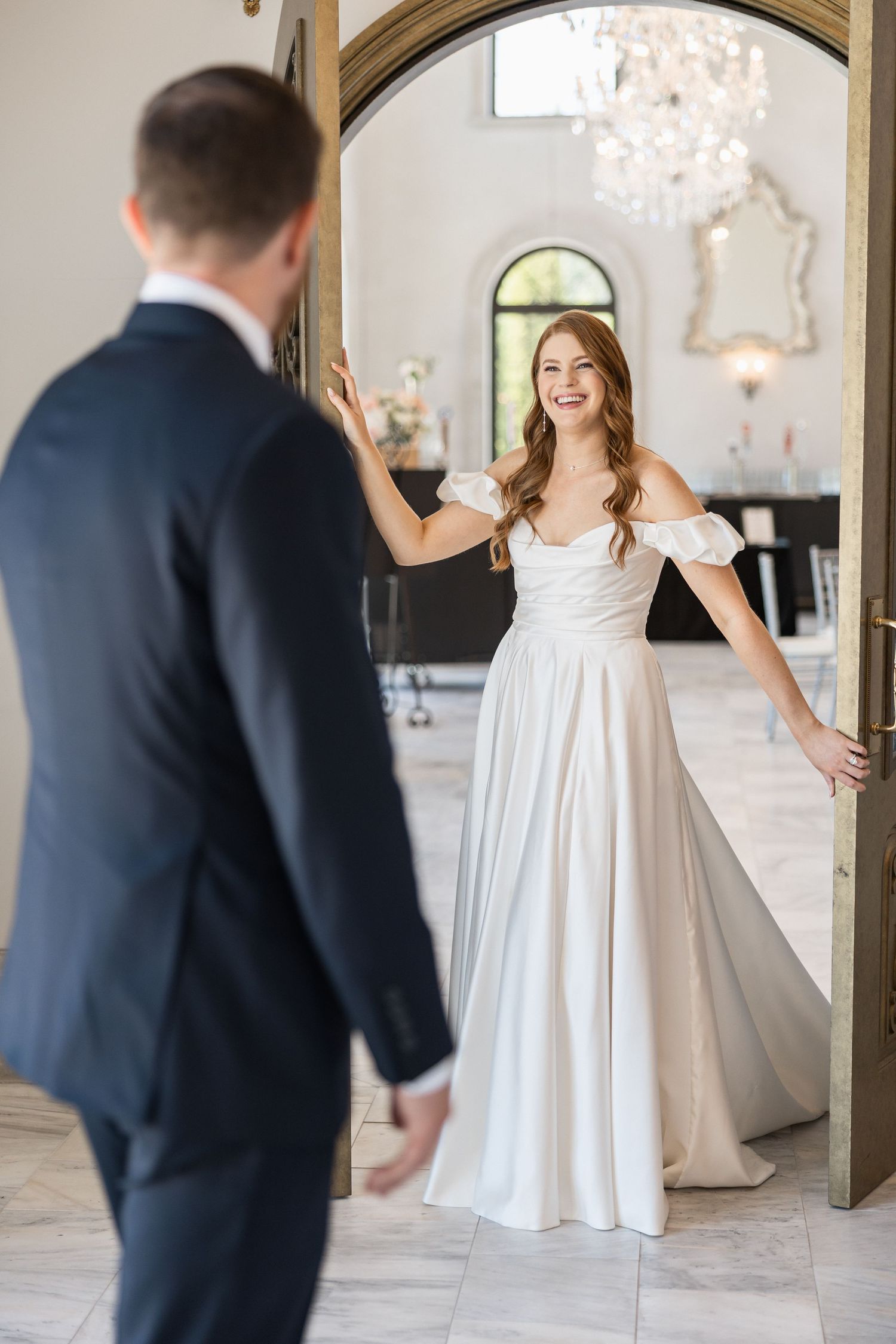 Bride in flowing white gown reveals herself to groom in elegant indoor setting.