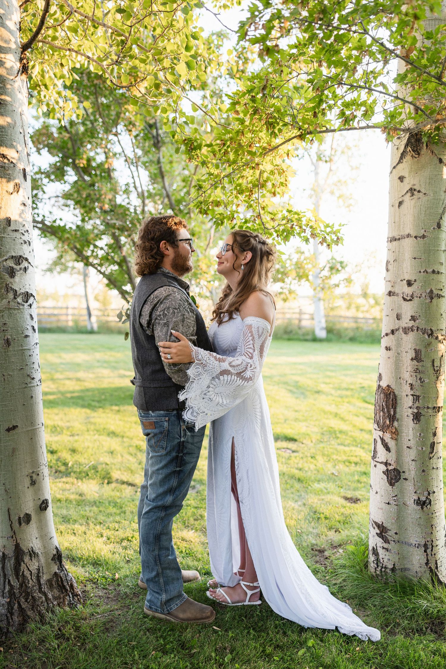 A romantic wedding couple embraces among white birch trees in a sunlit meadow during golden hour.