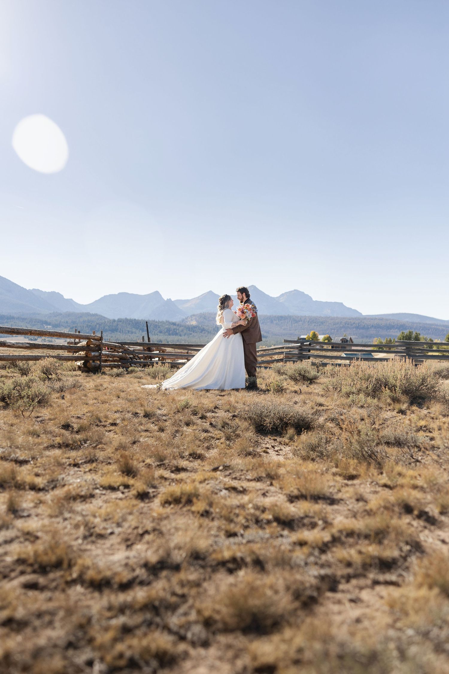 A couple in wedding attire stands together against a rustic mountain landscape with wooden fences and dry grasslands.